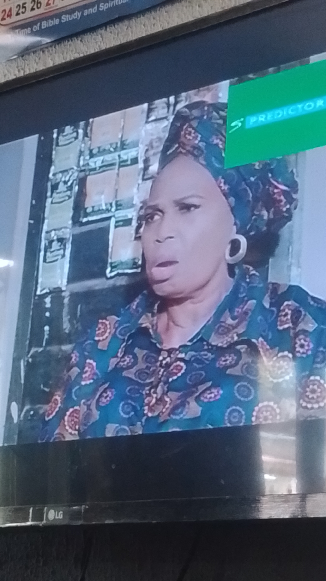 A close-up, medium shot captures an older Black woman from the chest up, looking to her left with her mouth open and a surprised or distressed expression. She wears a blue headscarf with a floral pattern in pink, white, and brown, and a matching blue collared shirt adorned with a similar floral design. Large, circular earrings are visible on her earlobes. Behind her, a wall is covered with what appear to be numerous small, beige packets of items, possibly food or medication, arranged in vertical columns. A green overlay in the upper right corner of the image displays the white text "S PREDICTOR". The bottom of the image shows the LG logo on a black television frame.