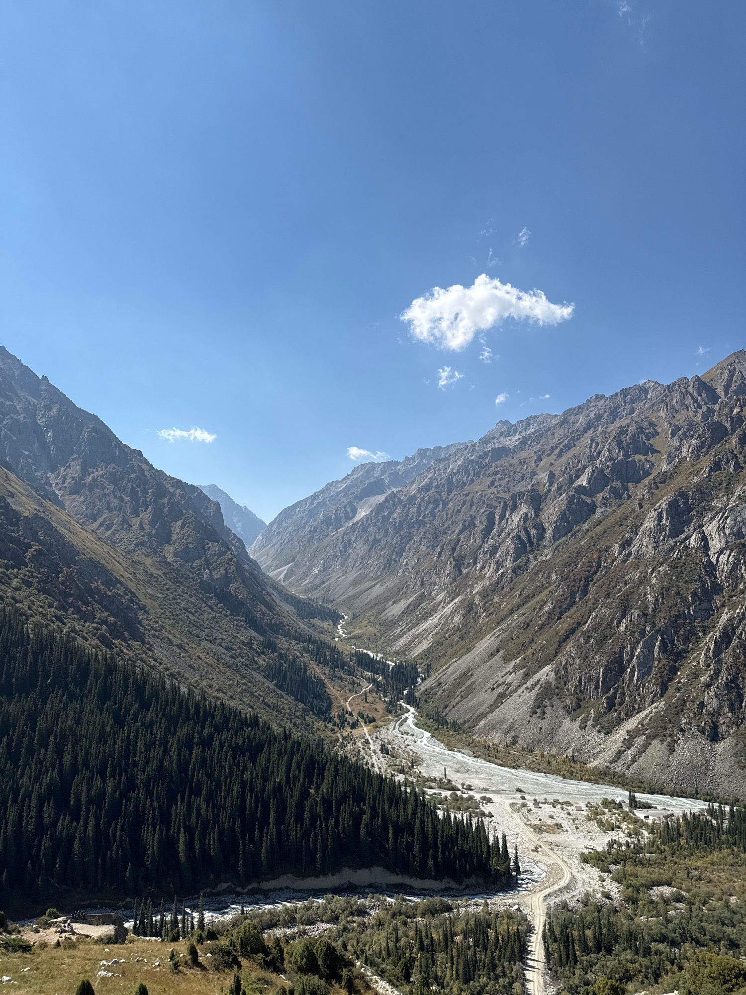 This is a high-angle, wide shot of a mountain valley in Kyrgyzstan. The valley is narrow, with steep, rocky slopes on either side. A river flows through the bottom of the valley, and there is a dirt road that runs alongside it. The slopes of the mountains are covered with trees in some areas, and in other areas, they are barren rock. The sky is blue with a few white clouds. The overall impression is one of a remote and rugged natural landscape.
