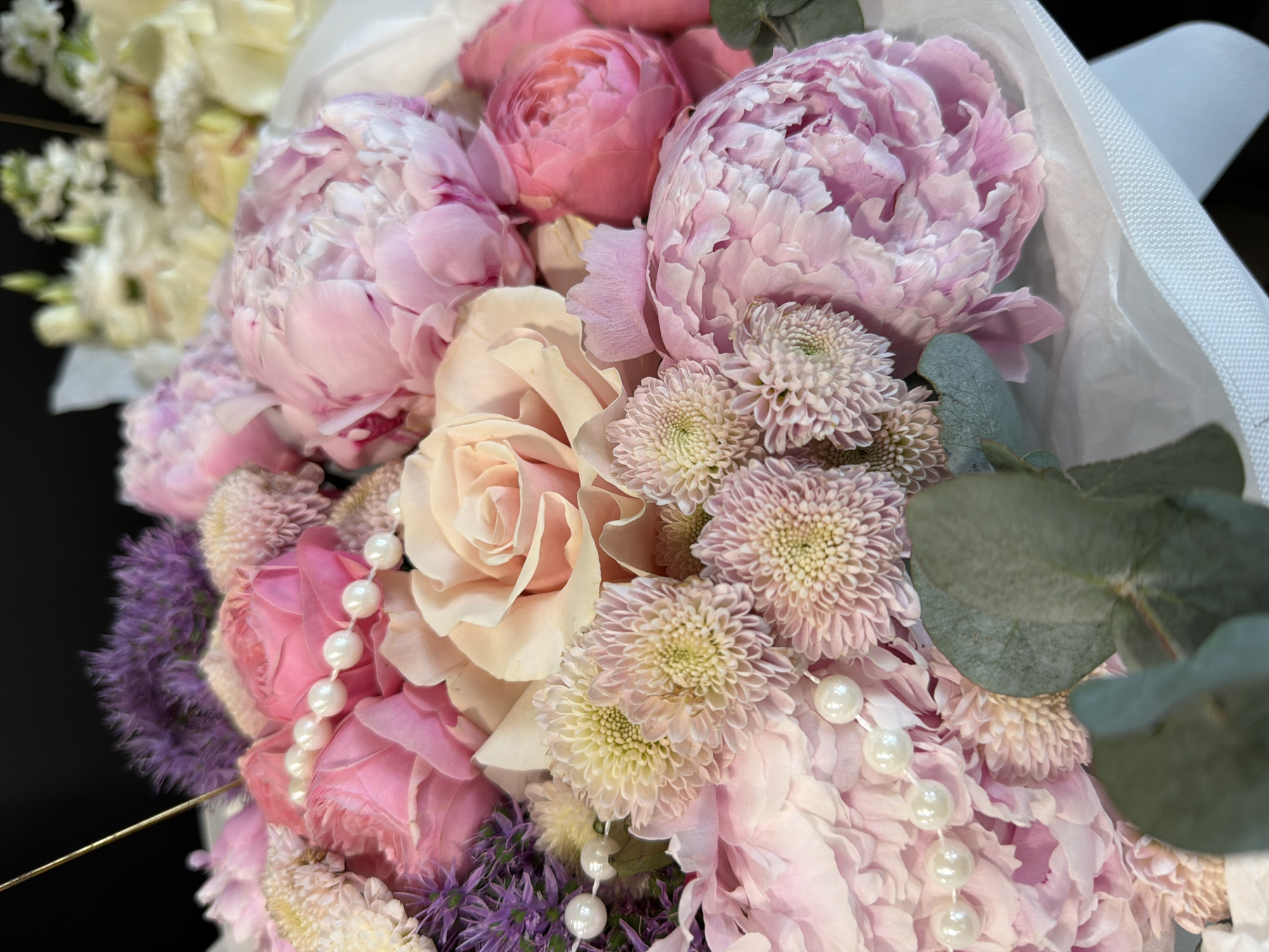 This is a close-up, eye-level shot of a lush floral arrangement. The bouquet is composed of various flowers in shades of pink, cream, and purple, including peonies, roses, chrysanthemums, and possibly some filler flowers. Several strands of pearl beads are incorporated into the arrangement, adding a touch of elegance. There are also some green eucalyptus leaves visible, adding texture and contrast. The flowers appear to be freshly cut and are artfully arranged, suggesting they are part of a bouquet for a special occasion. The background is dark and out of focus, drawing full attention to the intricate details and soft textures of the flowers. The lighting is soft and diffused, highlighting the delicate petals and subtle color variations. There is no visible text in the image.