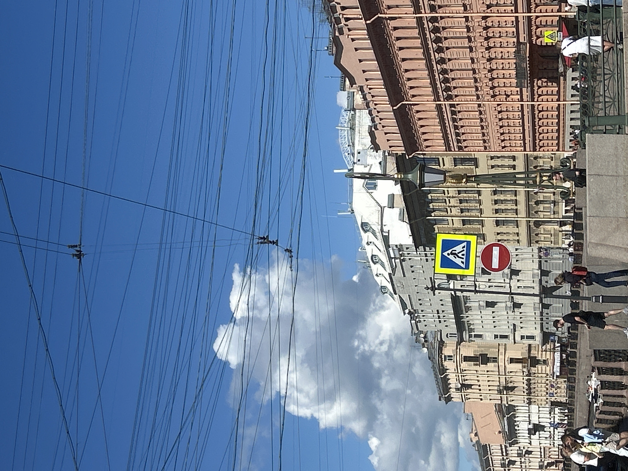 The image depicts a street scene in a city. A clear blue sky with scattered white clouds serves as the backdrop, overlaid by a dense network of overhead wires. In the middle ground, several people are walking along a sidewalk next to a canal or river. Buildings with traditional architecture, featuring ornate facades and multiple windows, line the street. Traffic signs, including a pedestrian crossing sign and a "no entry" sign, are visible on poles. In the foreground, people are visible on the sidewalk, and a decorative lamp post stands prominently. The overall impression is of a bustling urban environment on a sunny day.