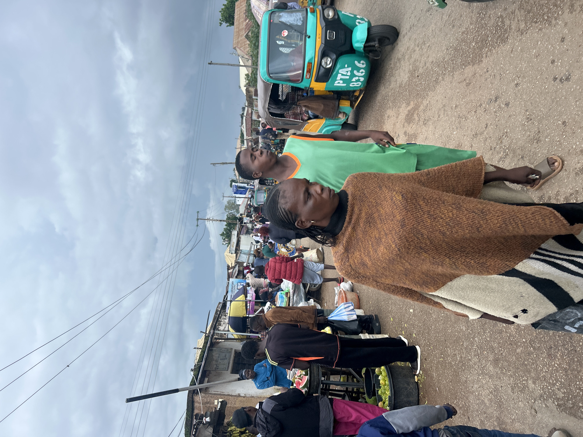 The image depicts a busy street market scene in Jos, Nigeria, under a cloudy sky. In the foreground, a woman wearing a brown knitted poncho over a patterned garment walks towards the left, while a young man in a bright green sleeveless shirt walks beside her, looking down. Behind them, a teal and yellow auto-rickshaw, a common mode of transport in the region, is parked, displaying the license plate "PTA-8366". The market is lined with stalls displaying various goods, including fruits like watermelons and what appear to be limes or lemons in large black buckets. Several people are seen browsing and interacting at the stalls, while others walk through the crowded street. The overall environment suggests a lively and active marketplace.