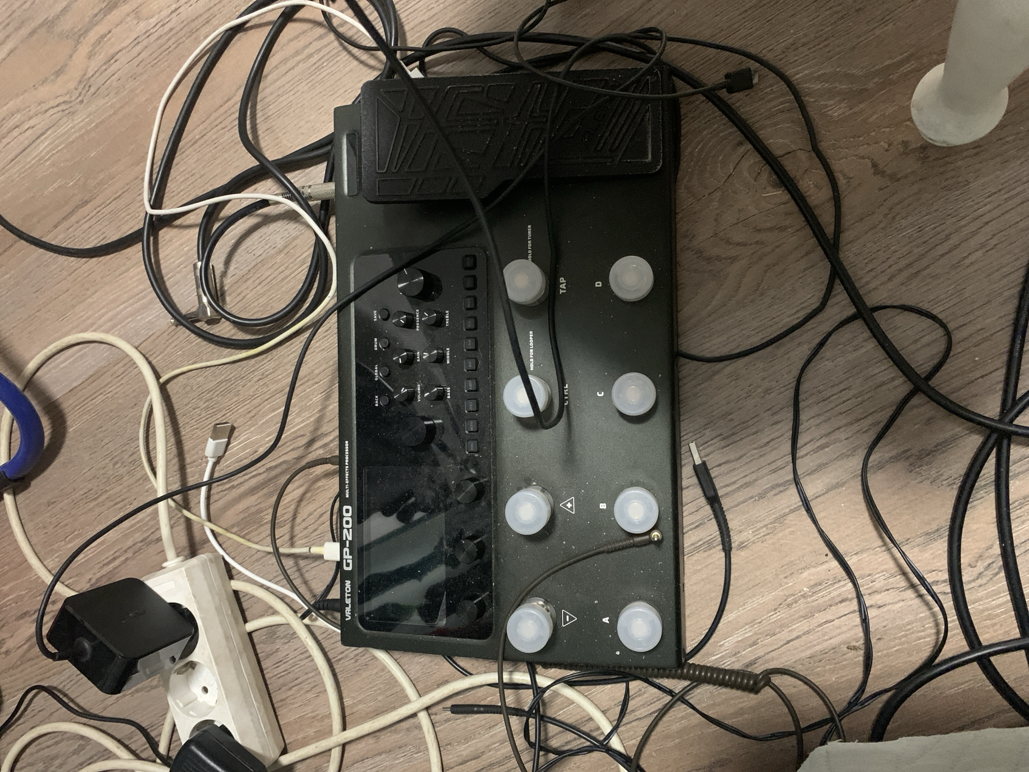 The image shows a Valeton GP-200 multi-effects processor lying on a wooden floor, entangled with various cables and power adapters. A white power strip with a black adapter plugged into it is visible in the upper left corner. The processor itself has a screen, several knobs, buttons, and footswitches. Some of the labels visible on the processor include "VALETON GP-200", "MULTI-EFFECTS PROCESSOR", "BACK", "GLOBAL", "DRUM", "SAVE", "VOLUME", "GAIN", "PRESENCE", "BASS", "MIDDLE", "TREBLE", "AMP", "CAB", "MOD", "DELAY", "REVERB", "HOLD FOR LOOPER", "HOLD FOR TUNER", "TAP", and buttons labeled "A", "B", "C", and "D". The scene appears to be an indoor environment, likely a room, with the wooden floor being the primary visible surface. There are no people present in the image. The image is a close-up shot, focusing on the electronic equipment and its surrounding cables. The location is identified as Nagayevo, Russia.