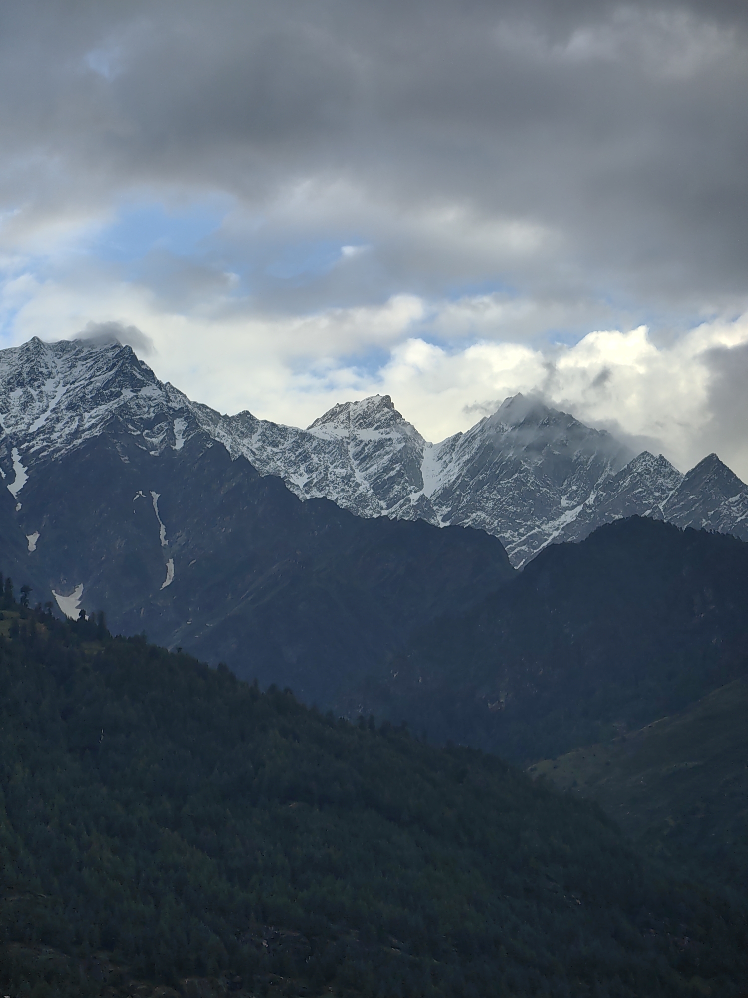 The image depicts a panoramic view of snow-capped mountains in the city of Vashisht, India. The foreground is dominated by a dense forest of dark green trees, which gradually recedes into the darker, shadowed slopes of the mountains. The middle ground features the imposing, rugged peaks of the mountains, their upper reaches covered in snow. The snow appears patchy, revealing dark rock formations underneath. The sky above is a dramatic mix of dark, heavy clouds and patches of lighter blue sky, with some white clouds interspersed. The lighting suggests an overcast or post-storm condition, casting a somber mood over the landscape. There are no people or animals visible in the frame. The scene is a natural, mountainous environment, characterized by its altitude and weather.