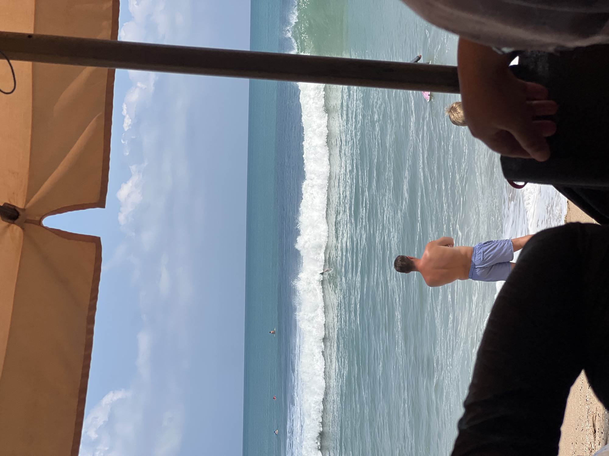 The image shows a beach scene in Kuta, Indonesia, viewed from under an umbrella. In the foreground, partially visible arms and hands suggest people are sitting or reclining. The middle ground features a beach with a man standing at the water's edge, facing the ocean, wearing light blue patterned swim trunks. In the ocean, several surfers are visible, with one actively riding a wave. The water is turquoise and calm, with a moderate surf break. The sky is blue with scattered white clouds.