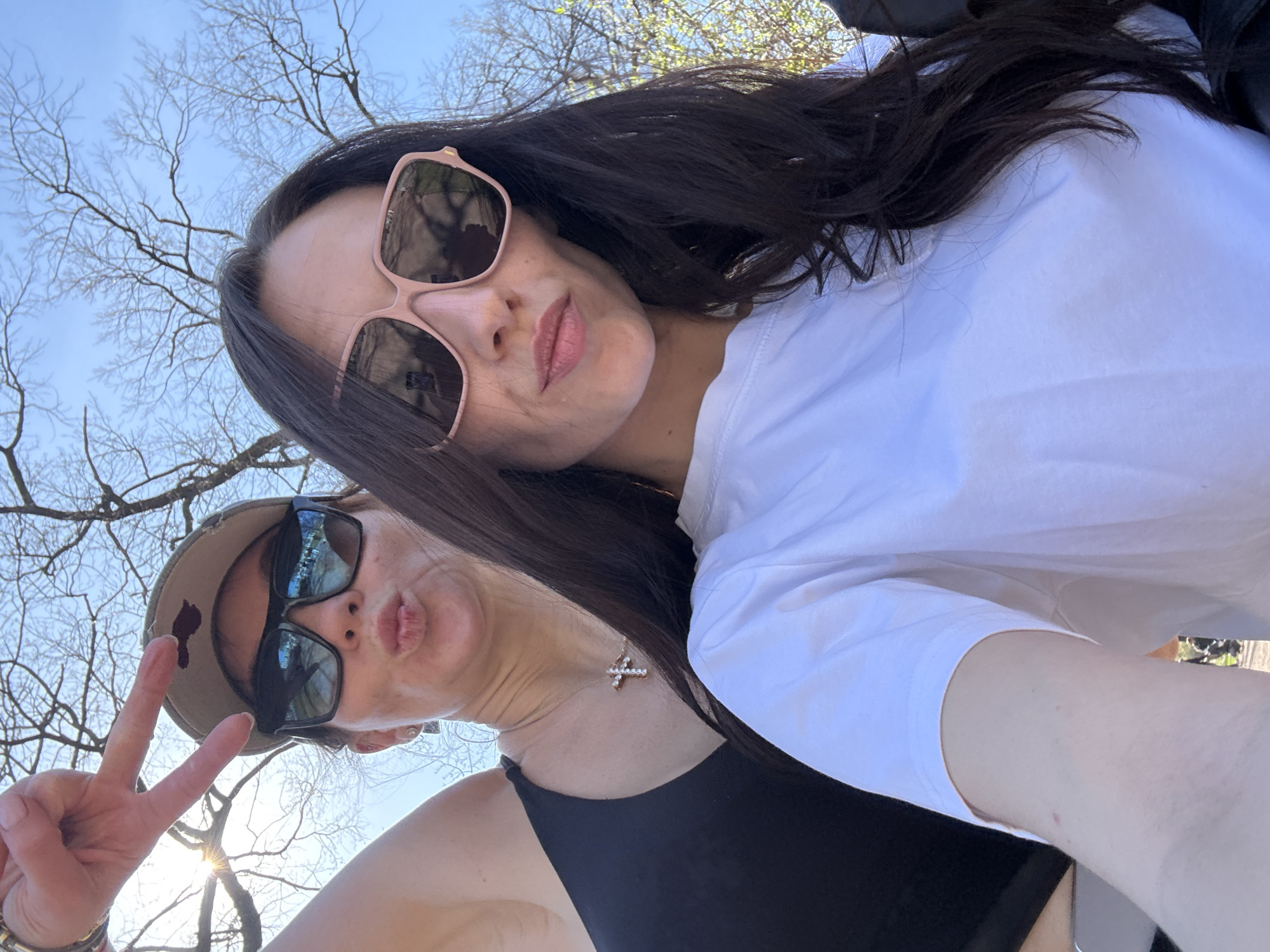 Two women are taking a selfie outdoors. The woman in the foreground wears a black tank top, a cap, and sunglasses and is making a peace sign with her hand while pouting her lips. The woman in the background wears a white shirt and pink-framed sunglasses. Bare trees and a blue sky are visible in the background. The location appears to be outdoors, possibly in a park, though there is insufficient information to determine whether it is Saint Petersburg, Russia.