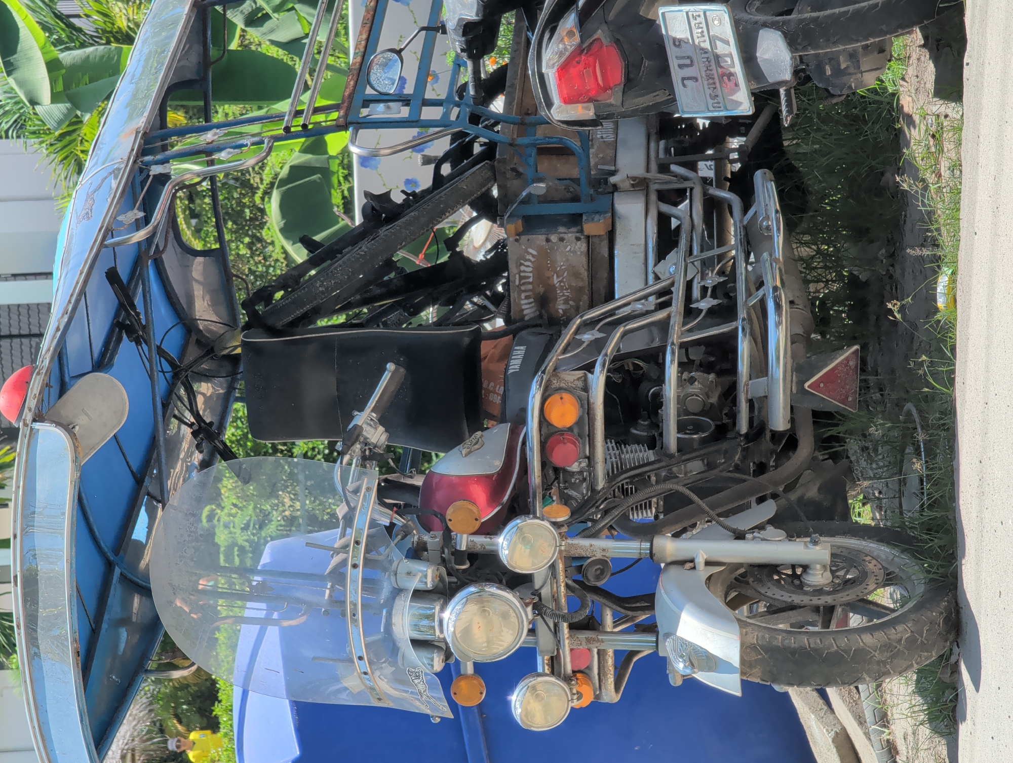 The image shows a parked tuk-tuk in ตำบลเกาะพะงัน, Thailand. The vehicle features a motorcycle front end with multiple headlights and a blue passenger compartment. The rear section includes a cargo area with metallic framework. A license plate is partially visible. The vehicle is parked on the edge of a concrete surface adjacent to grassy vegetation.