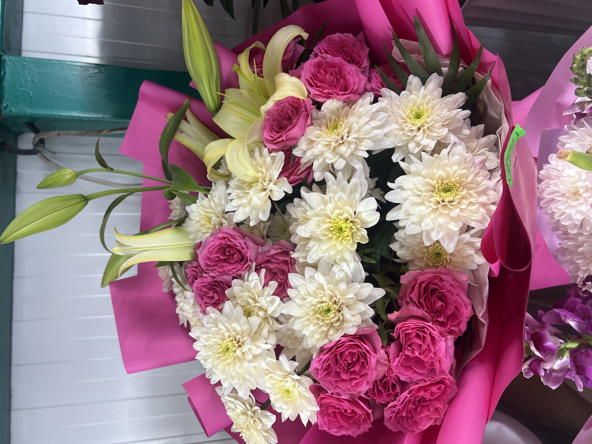 This is a close-up, eye-level, medium shot of a vibrantly colored flower bouquet. The bouquet is predominantly composed of pink roses and white chrysanthemums, with accents of yellow lilies and green foliage. The flowers are arranged in a lush, full bouquet, wrapped in bright pink paper that forms a dramatic backdrop. The bouquet is set against a white, vertically paneled background, and a section of a dark green, industrial-looking structure is visible in the upper right corner. The lighting is bright, highlighting the texture and colors of the flowers. There are no people or animals visible in the image, nor any specific landmarks that would definitively place it in Chișinău, Moldova.