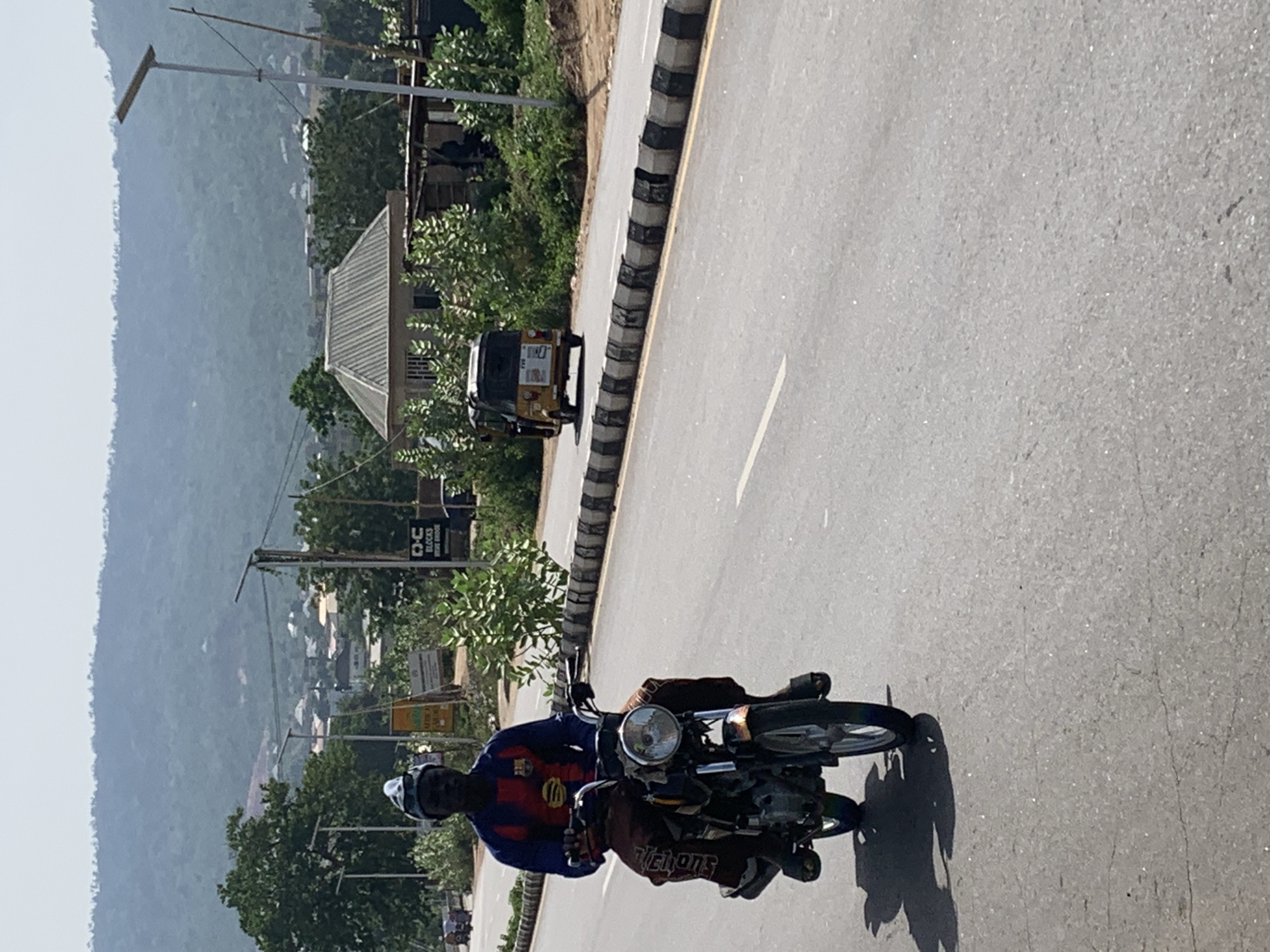 A man on a motorcycle is riding on a paved road in Zango-ndaji, Nigeria. The man is wearing a helmet and a blue and red shirt. In the background, there is a three-wheeled taxi and a hill.