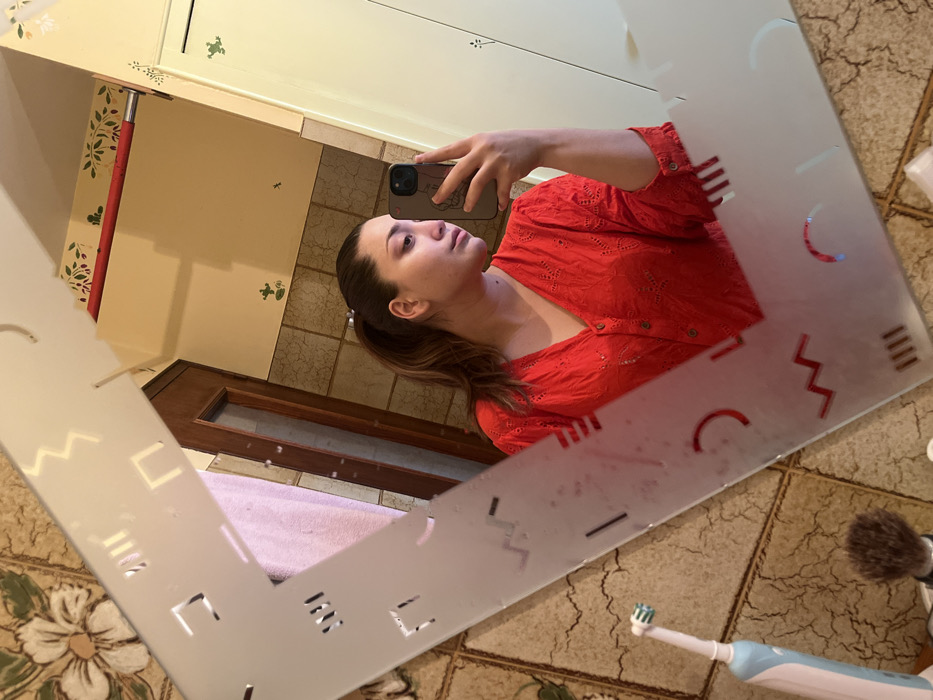 The image displays a female individual taking a mirror selfie in a bathroom, identified as being in Ancenis, France. The subject is reflected from the chest up, holding a dark-colored smartphone in her left hand (appearing as her right in the reflection), positioned to capture the image. She has light brown hair styled in a ponytail and is wearing a red, long-sleeved, V-neck top with front buttons and eyelet embroidery.
The mirror features a frosted frame with geometric cutouts including zigzags, straight lines, rectangles, and semicircles; small water droplets are present on the mirror's surface. The reflected background reveals a bathroom interior: the lower wall section is covered with brown, textured square tiles, while the upper wall is painted a light cream color. Decorative elements on the upper wall include a red border strip with a leaf pattern and several small green frog decals.
On a surface to the left of the subject's reflection, a light blue and white electric toothbrush and a shaving brush with a dark handle are visible. Further within the reflection, a dark wooden door with a vertical glass panel and a light pink towel are discernible. A light-colored cabinet or door is visible to the right of the subject's reflection.