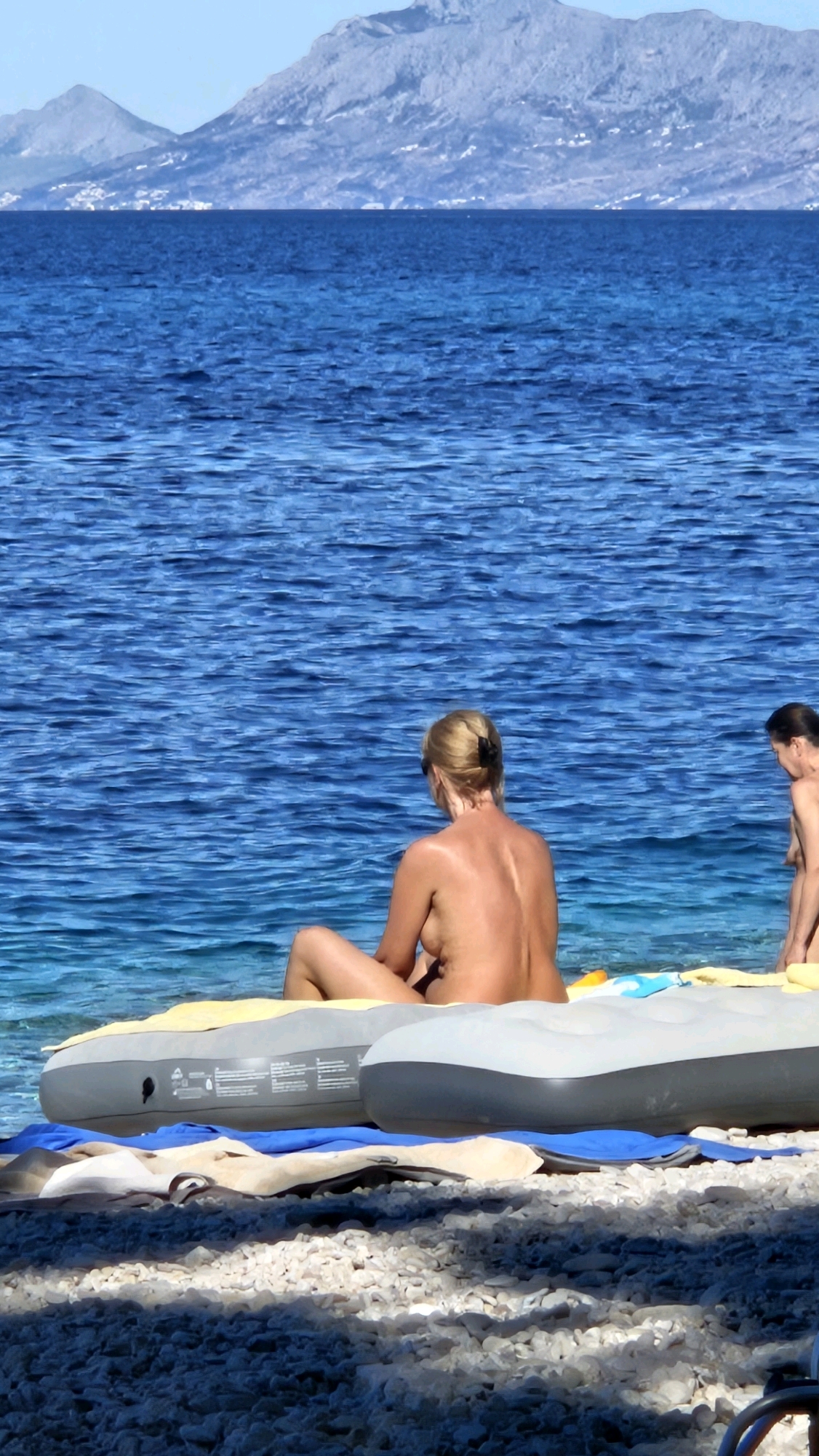 In Općina Podgora, Croatia, two nude individuals are relaxing on a pebble beach. In the foreground, a blonde woman with her back to the camera sits on an inflatable mattress and towels. To her right, another individual, whose face is not visible, also appears to be nude and is partially obscured. The sea, a vibrant blue, stretches out behind them, meeting a distant, hazy mountain range under a clear sky. The beach itself is covered in small, white pebbles, with dappled sunlight creating shadows.