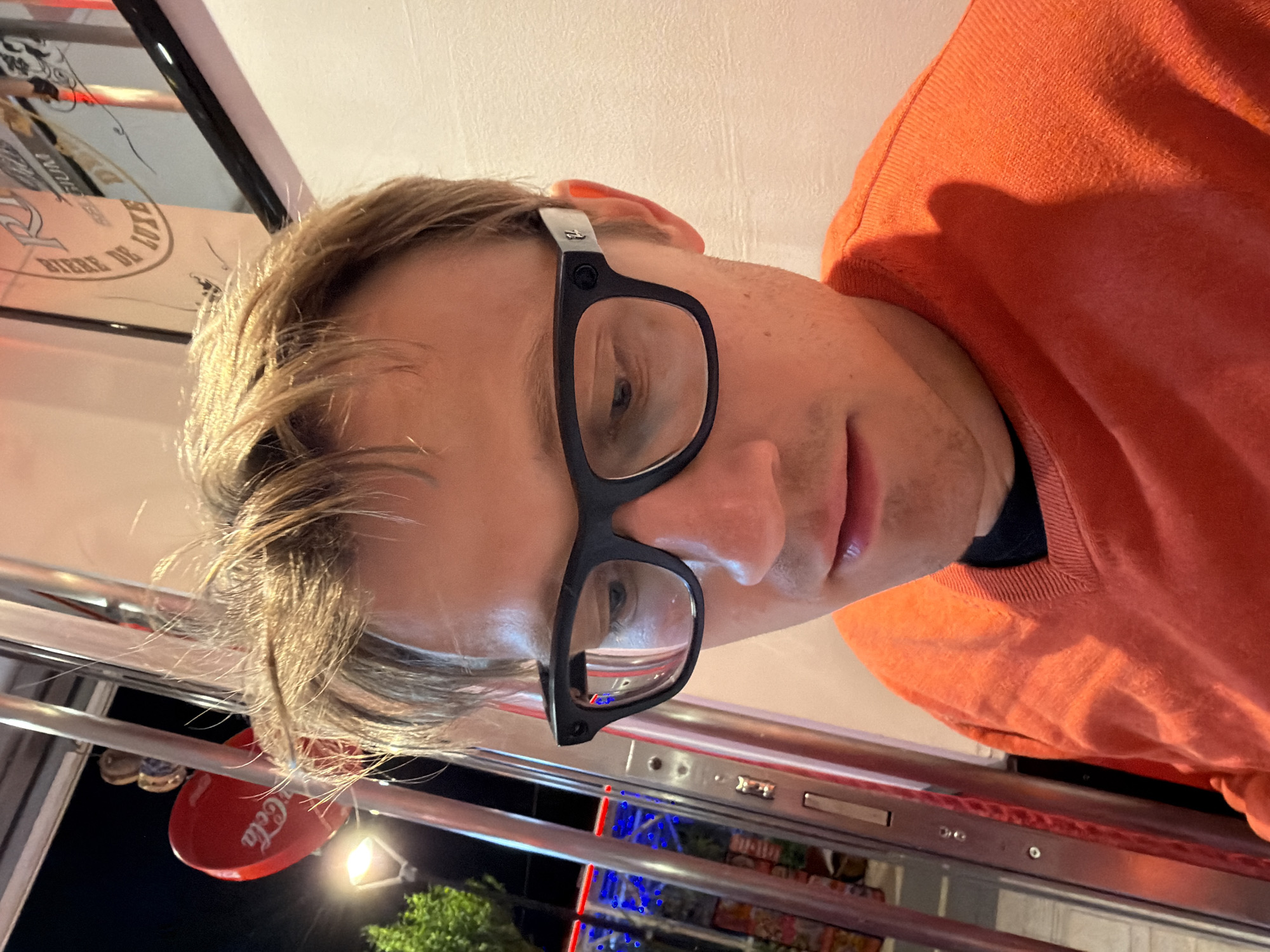 A selfie, rotated, shows a man with blond hair, wearing black-rimmed glasses and an orange sweater. He is seated at a table with a silver metal frame, possibly in a diner or restaurant. The table is partially visible, with a red Coca-Cola branded object hanging nearby. Through a window, there's a partial view of a tree and night sky, with lights suggesting an urban environment. Based on contextual clues, the location is Ragusa, Italy. A framed image on the wall is partially visible.