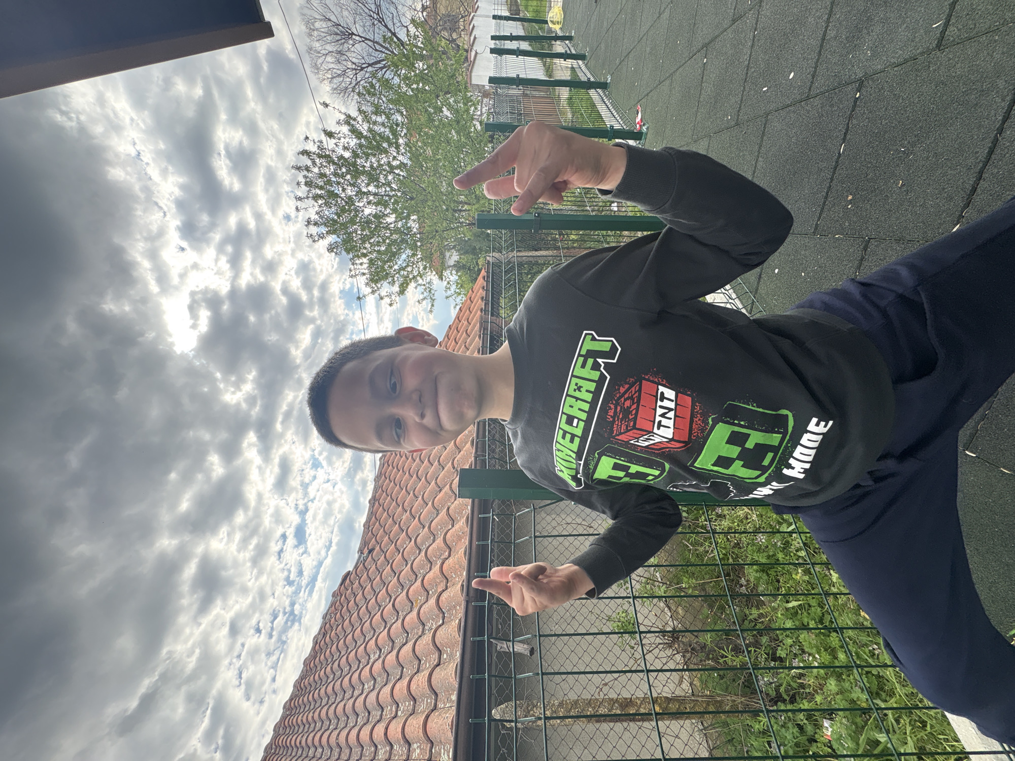 A person stands on a playground or similar recreation area with a green and grey tiled surface. The person, likely a young male, wears a dark long-sleeved shirt with a Minecraft design and dark-colored pants. They are making a hand gesture. Behind the person is a green metal fence and vegetation. In the background, there's a building with a red tiled roof, a tree with light green leaves, and a sky filled with clouds. The location is an urban area of Unknown, Unknown.