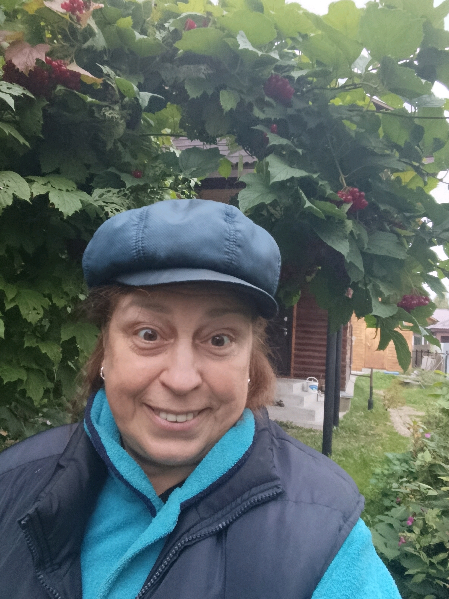 A middle-aged woman wearing a blue cap, a dark vest, and a bright blue scarf smiles at the camera, with her eyes wide open and her head slightly tilted. She is standing outdoors in front of a building with wooden siding and steps. Behind her, there are green leafy branches with red berries. The environment appears to be a residential area in Veriya, Russia, with greenery and a rustic building suggesting a rural or suburban setting.