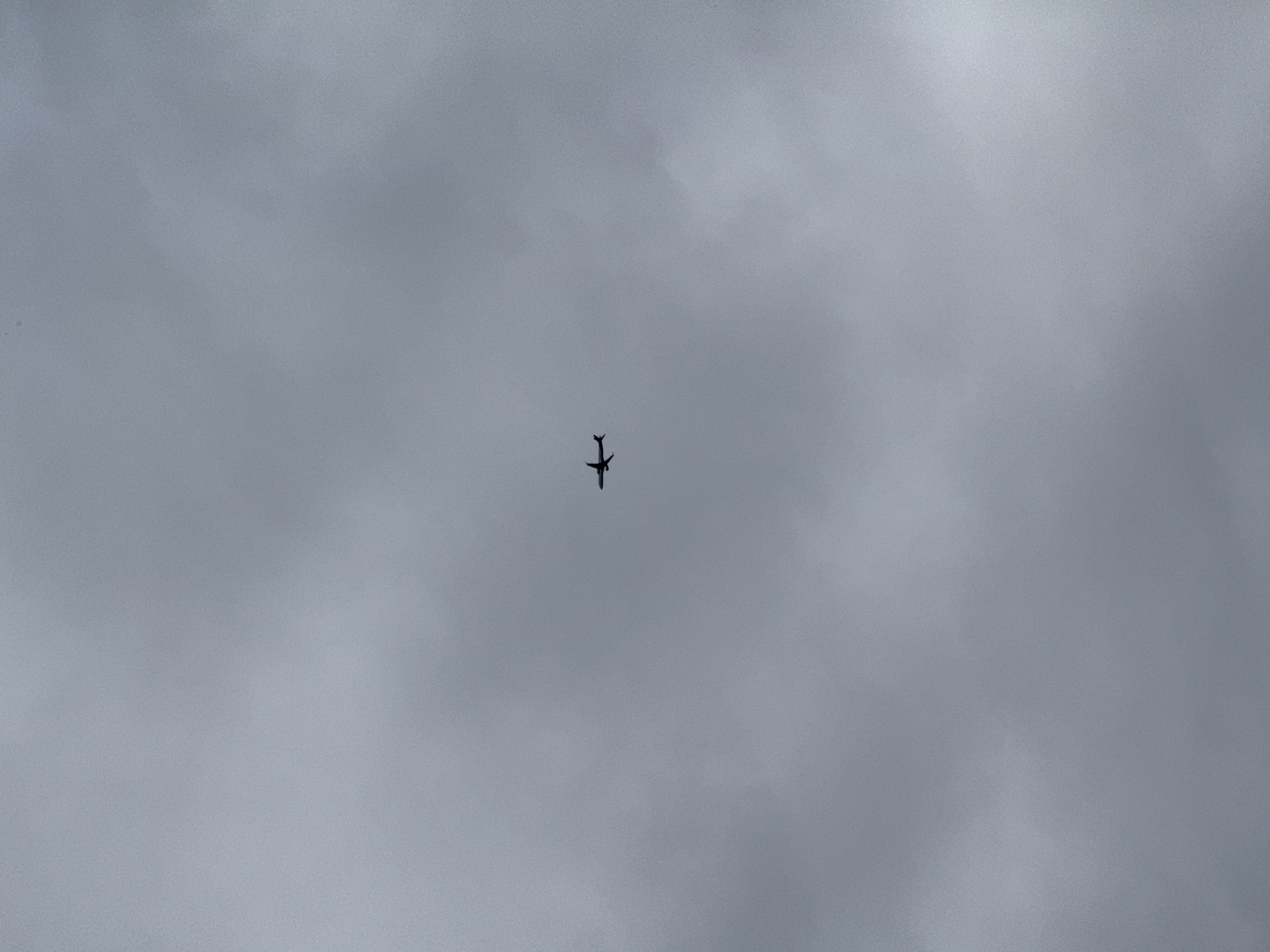 An aircraft is flying through an overcast sky. The photograph appears to be taken from the ground, looking upwards towards the plane. The background is uniformly gray and featureless, indicating a cloudy or overcast day. The aircraft is a dark silhouette against the gray sky. The location is Løvstad, Norway.