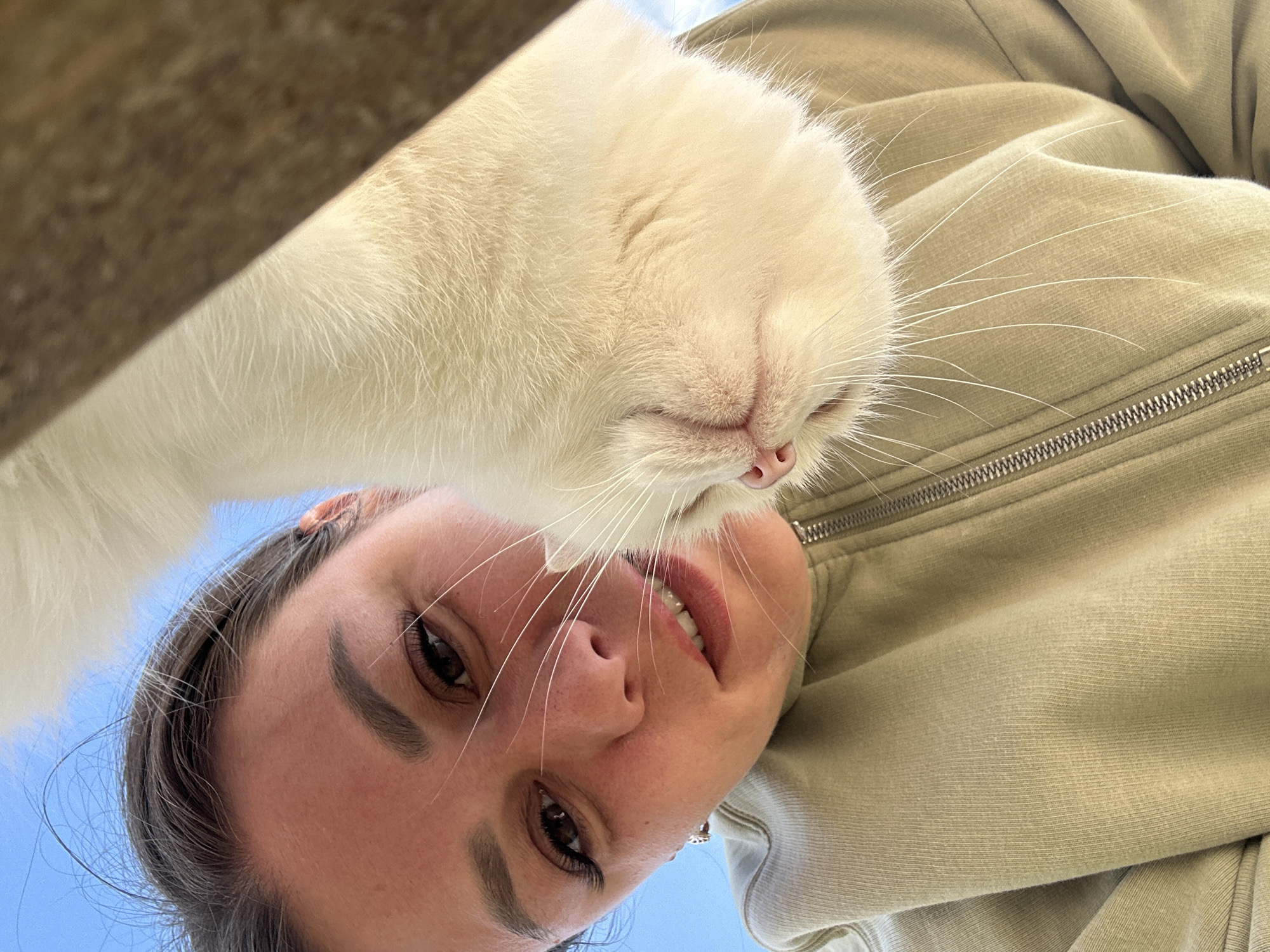 A low-angle, close-up shot captures a young woman's face and a white cat's head in the foreground, set against a bright blue sky and a wooden beam. The woman, with her dark hair pulled back and wearing a light green zip-up hoodie, looks directly at the camera with her mouth slightly open, revealing her teeth. Her right eye is visible, accentuated by long eyelashes. The cat's white fur dominates the right side of the frame, with its pink nose and long white whiskers prominent. The image appears to be a selfie, possibly taken outdoors on a sunny day. There is no indication of the specific location within the city of Chaikovsky, Russia, nor any context to suggest the activity beyond a casual interaction between a person and a pet.