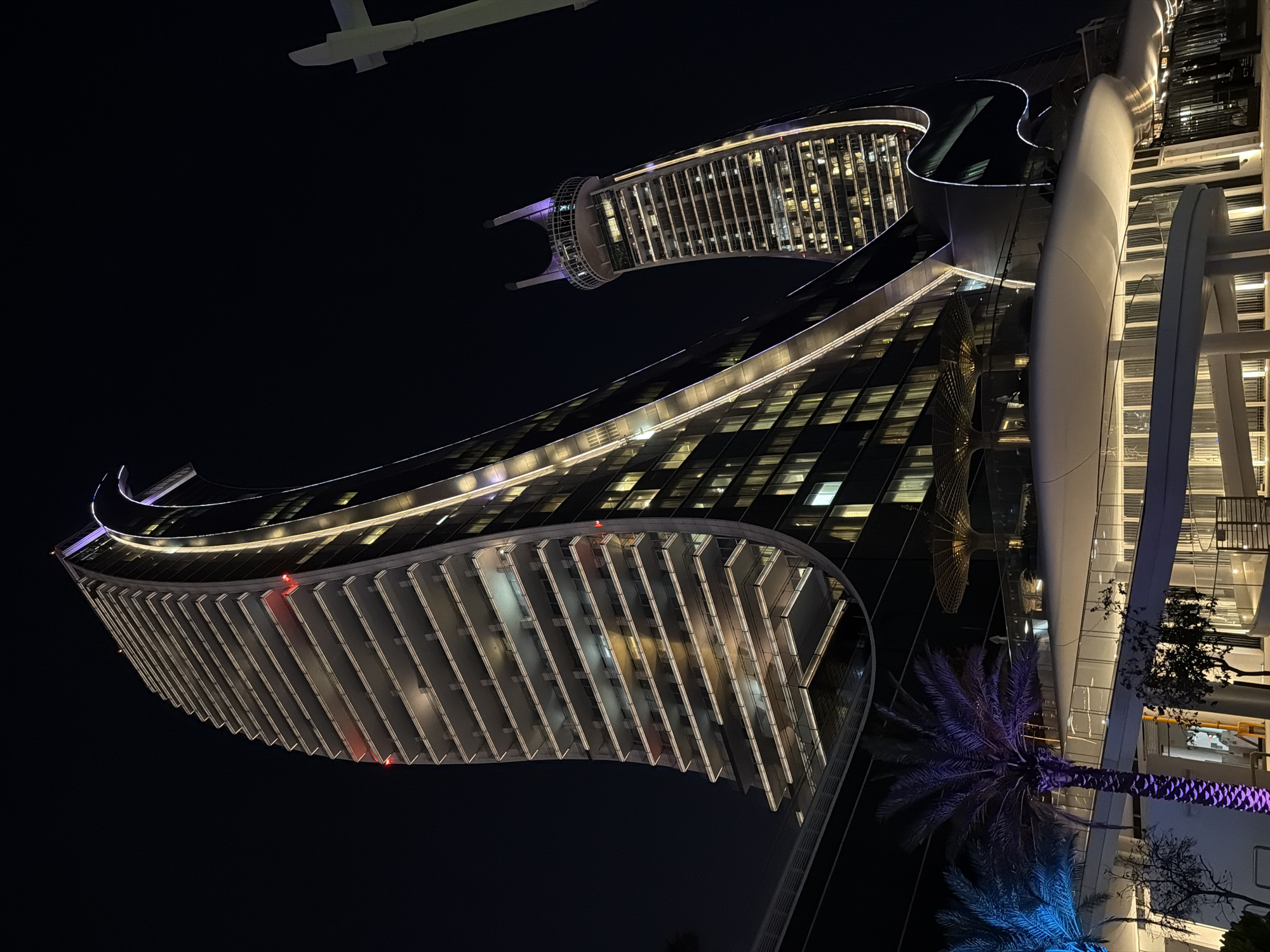 A wide-angle, low-angle, night exterior shot shows a modern architectural complex with two distinct buildings. The building in the foreground is a tall, curved skyscraper with numerous balconies and white horizontal accents. Its facade is primarily dark glass, with vertical lines of light tracing its curvature. The building on the right is also a skyscraper, but it is more slender and features a distinctive top with two curved protrusions. It is illuminated with warm interior lights visible through its windows.
In the lower portion of the image, the base of the foreground building is visible, with a white, organic-shaped structure that appears to be a terrace or walkway. Palm trees with purple and blue lighting are present to the left, adding a splash of color to the scene. The sky is dark, indicating nighttime. A white, T-shaped structure is partially visible in the upper right corner, possibly a lamppost or part of another building. The overall impression is one of a luxurious, contemporary urban environment.