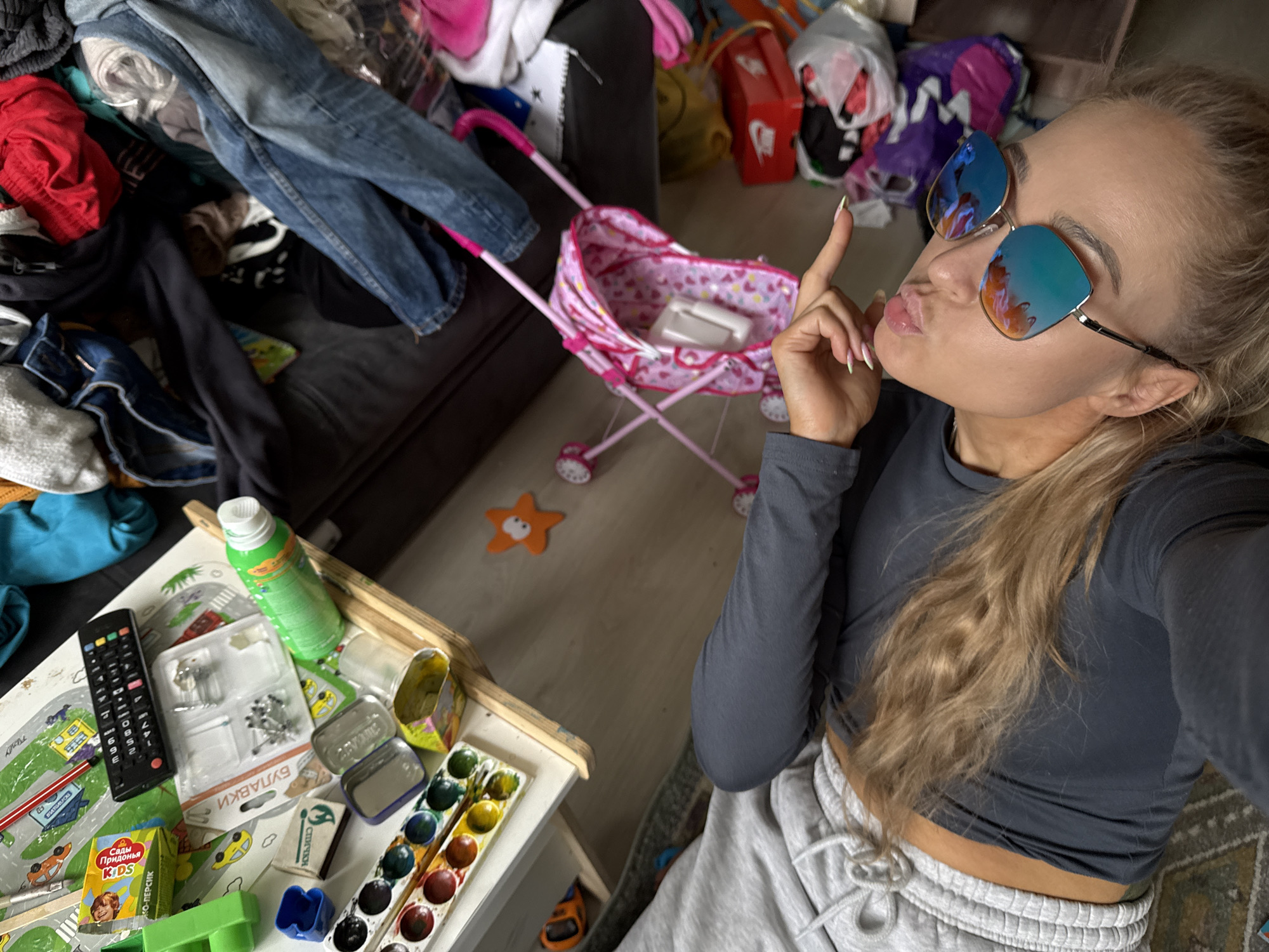 A young woman with long, light-brown or blonde hair, pulled back into a ponytail, takes a high-angle selfie in a cluttered indoor environment. She wears reflective blue and orange sunglasses, a dark grey cropped long-sleeved top, and light grey sweatpants. Her lips are pouted, and her right hand, featuring long fingernails with light-colored tips, is raised with an index finger pointing upwards, indicating a playful or sassy pose.
The setting is a room, likely a living area or bedroom, characterized by significant disarray and numerous household items. In the immediate foreground, on a white table covered with a road-themed playmat, rests a black remote control, a green spray bottle, a package of thumbtacks labeled "БУЛАВКИ" (PINS) and "25ШТ" (25 PCS), an open tin can, a watercolor paint palette, a matchbox with "СПИЧКИ" (MATCHES) written on it, and a "Сады Придонья KIDS" (Pridonie Gardens KIDS) juice box with "ЯБЛОКО-ПЕРСИК" (APPLE-PEACH) flavor. A green plastic toy is also visible on the table.
In the midground, a pink toy stroller adorned with heart patterns stands on a light-colored floor, containing a white object. An orange star-shaped mat or toy is on the floor nearby. The background is dominated by piles of clothing, including denim jeans and red shorts, heaped on and around a dark-colored couch. Various bags, one purple and another revealing a red box with a "Nike" logo, contribute to the cluttered appearance of the room. The visible Cyrillic text on items like the pins, juice box, and matchbox confirms the location context of Chaikovsky, Russia. The overall impression is one of a casual, lived-in domestic space, possibly with children present given the toys and children's products.