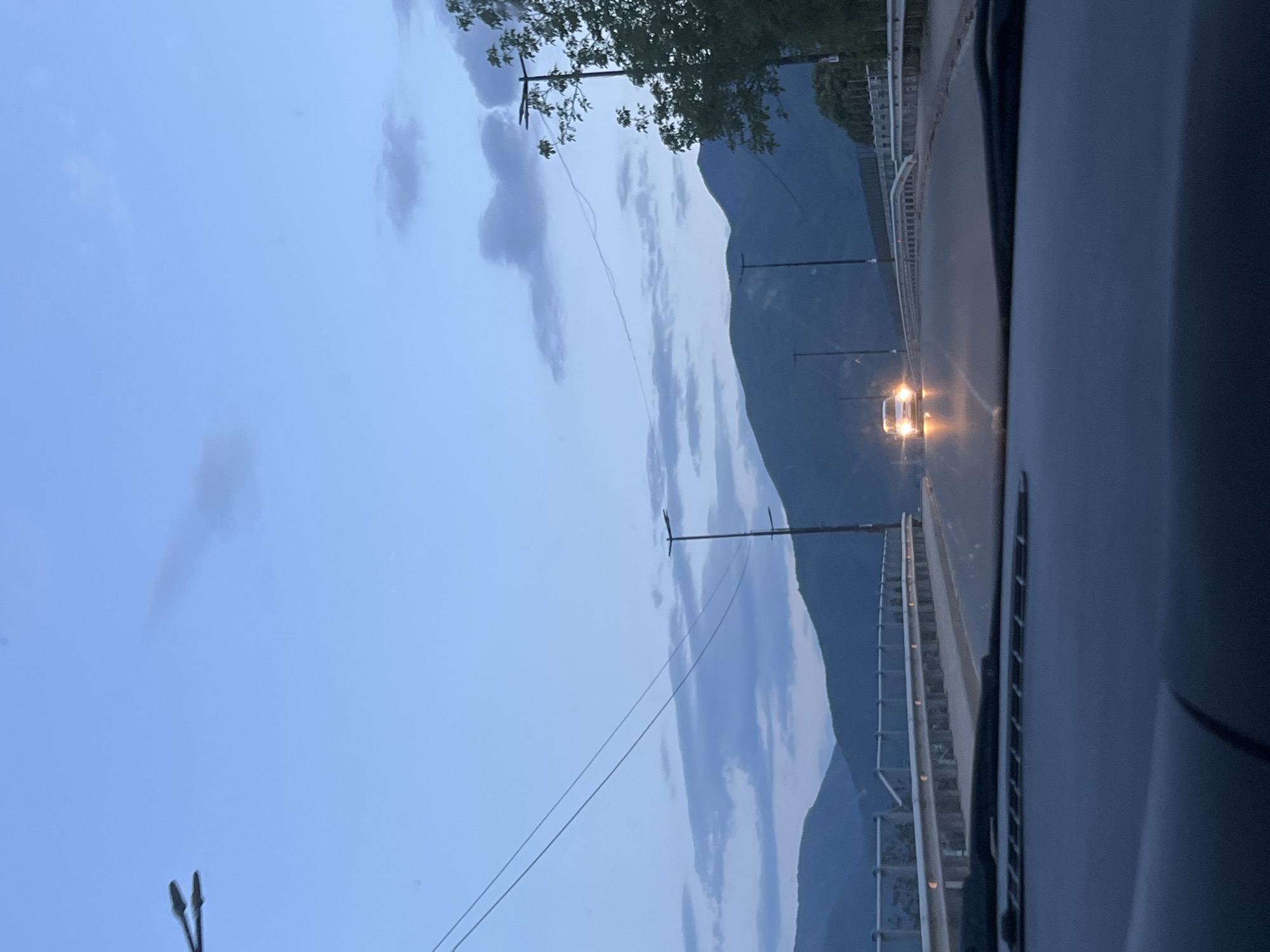 A photograph taken from inside a car shows a road or bridge at dusk or dawn with a car approaching. There is a mountain in the background with clouds in the sky. The location may be near Septemvri, Bulgaria, as the city is known for its surrounding mountains and roads. The road is lined with streetlights and a metal guardrail. Power lines stretch across the frame. A car's dashboard and part of the interior are visible, indicating the photo was taken from inside a vehicle. The approaching car's headlights are illuminated.