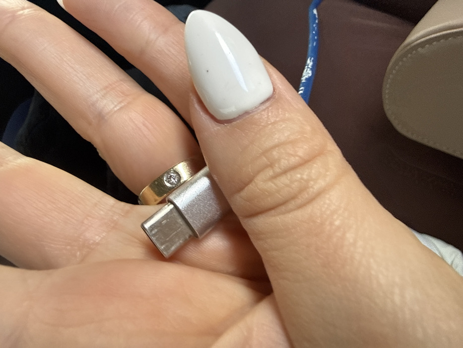An extreme close-up shot focuses on a person's left hand holding a silver-colored USB-C connector. The hand exhibits light-toned skin with visible wrinkles and knuckle lines. A gold-colored band ring, set with a single round clear stone, is visible on the ring finger. The thumb, positioned to the right, has an elongated, oval-shaped fingernail coated in opaque white polish, featuring a small dark speck on its surface. The USB-C connector is oriented vertically, held between the thumb and the base of the index or middle finger. In the lower right background, a blurred segment of a blue cable with white markings is visible, alongside a curved edge of a light-colored, possibly beige or cream-colored, object, which appears to be part of a shoe or bag. The immediate environment is dimly lit and out of focus, suggesting an indoor or vehicle setting. The specific location in Apnagyugh, Armenia, cannot be determined from the image.