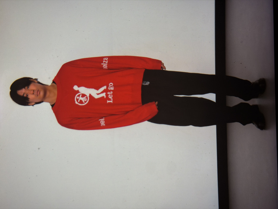 An indoor, full-body photograph depicts a male subject, appearing to be of East Asian descent, standing erect against a uniform off-white wall. The subject wears a red long-sleeved crew-neck sweatshirt featuring a white graphic on the chest: a stylized figure holding a circular object containing a stick figure, positioned directly above the white text "Let go." The left sleeve displays the vertical white text "ael.," and the right sleeve "elza." He is also dressed in loose black trousers and black dress shoes. His posture is frontal, with arms relaxed at his sides. The floor is a darker, likely gray, surface, demarcated from the wall by a distinct horizontal black line. The scene suggests a controlled studio environment. The specific location within London, United Kingdom, is not discernible from the provided imagery.