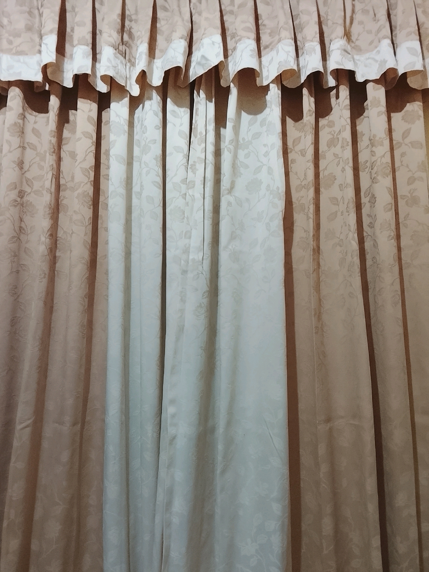 The image shows a close-up view of a set of closed curtains. The curtains are made of fabric with a floral pattern. The top portion of the curtains features a decorative valance with pleats and a contrasting lighter-colored trim. The main body of the curtains consists of draped fabric, with vertical folds that create a sense of depth and texture. There are two distinct color tones present in the curtains: a warmer, brownish-tan on the left and a lighter, creamier color on the right. The lighting casts shadows on the fabric, highlighting the texture and the folds. There are no people or other objects visible in the image, and the background is entirely composed of the curtains. The location of this image is within a dwelling in the city of Kalibeji, Indonesia.