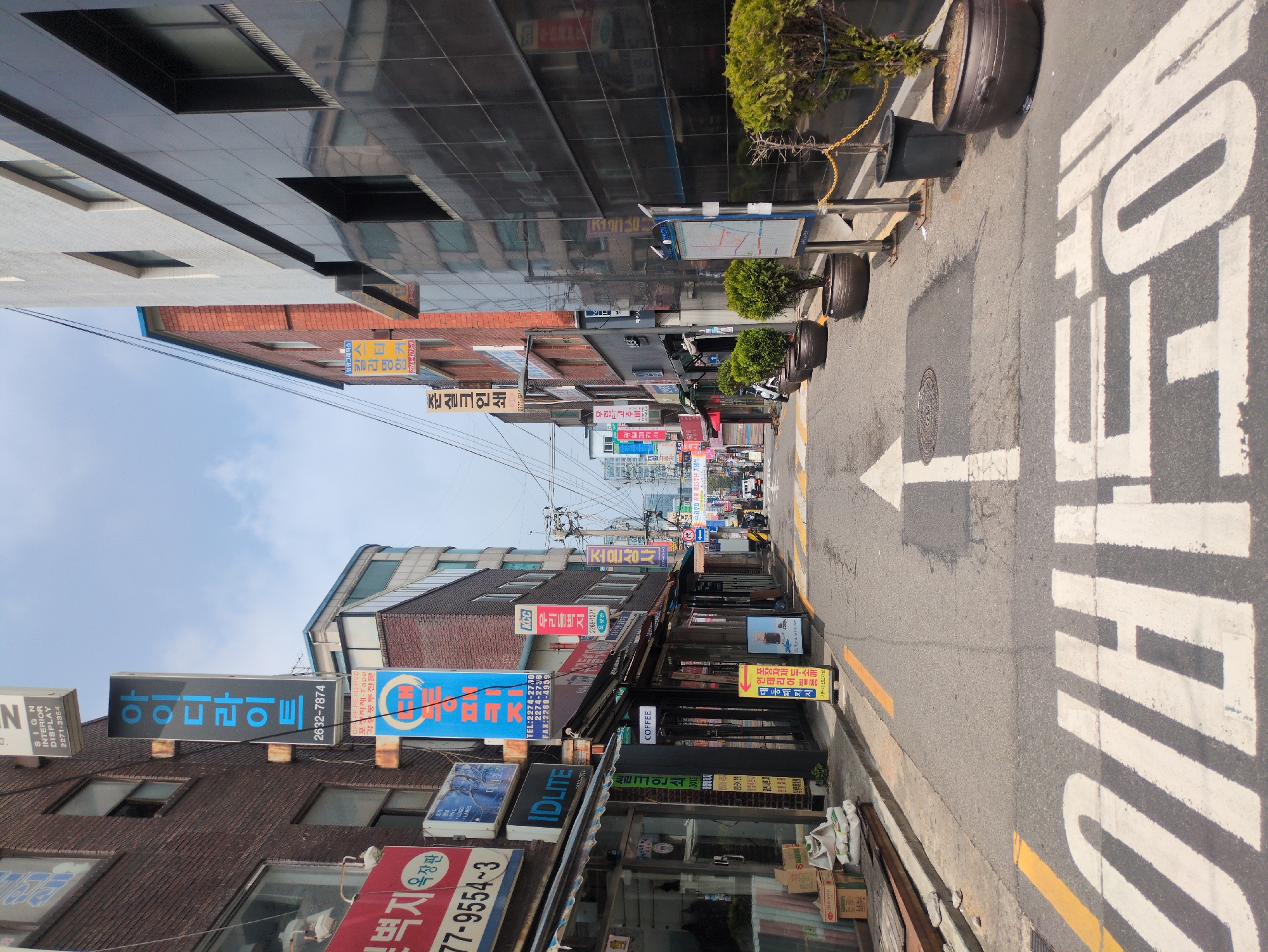 The image shows a narrow street in Seoul, South Korea, with buildings on both sides. Many Korean signs and storefronts line the street. A white directional arrow with Korean characters for "straight" is painted on the road. The street appears busy, with people and vehicles visible in the distance. Planters and some street furniture are visible on the right side of the street. Buildings exhibit a mix of architectural styles, including brick and modern glass facades. Power lines stretch across the sky.