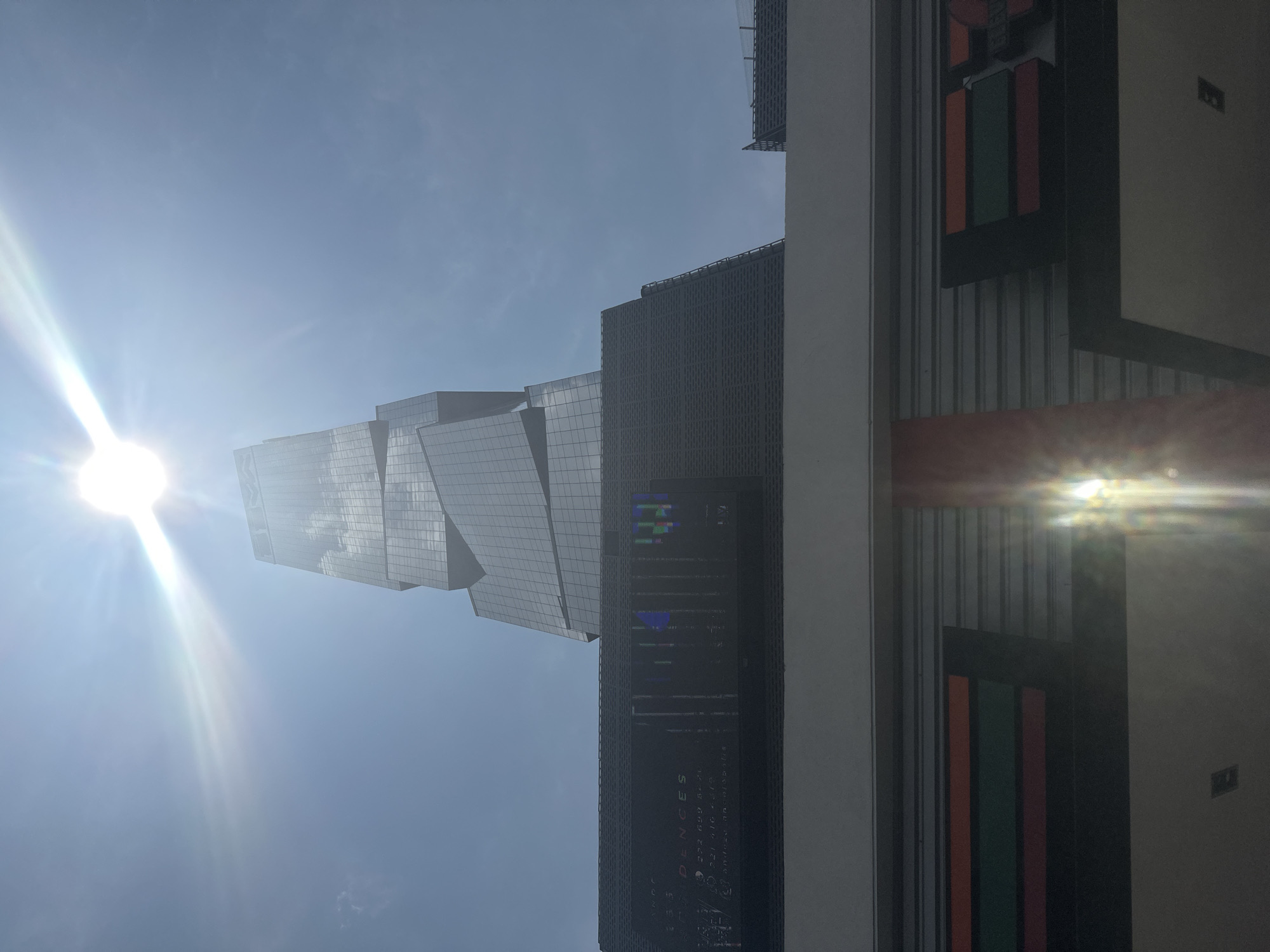 The image depicts an urban scene with modern architecture in San Bernardino Tlaxcalancingo, Mexico. In the foreground, a building with vertical striped accents of red and green is visible. A tall, contemporary building with a glass facade is prominent, reflecting the bright sunlight and surrounding sky. An electronic display on the side of the building shows various information. The sky is blue, and the sun is shining brightly, causing lens flare.