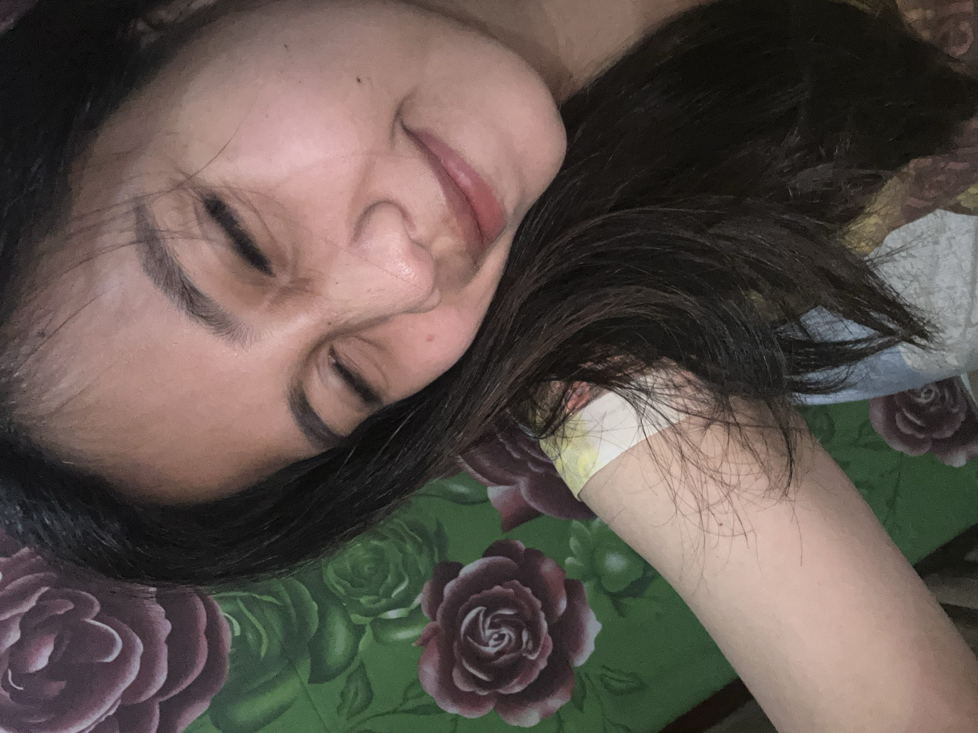 The media file shows a close-up, slightly angled, overhead shot of a person lying down with their eyes closed. The person has dark hair spread around their head and face, resting on a patterned surface. The surface appears to be a green fabric with a floral print, featuring large, stylized roses in shades of deep red and lighter pinks, with green leaves.
The person's face is turned to the side, with their cheek resting on the patterned surface. Their eyes are closed, and their lips are slightly parted, conveying a sense of relaxation or sleep. A portion of their arm is visible, resting near their head. There is a small, light-colored bandage or piece of tape on the inner wrist area of the arm. The lighting suggests an indoor setting, possibly at night or in a dimly lit room. There are no visible text elements in the image. The overall mood is peaceful and intimate. The location context of Depok, Indonesia, does not provide specific visual cues within the image itself.