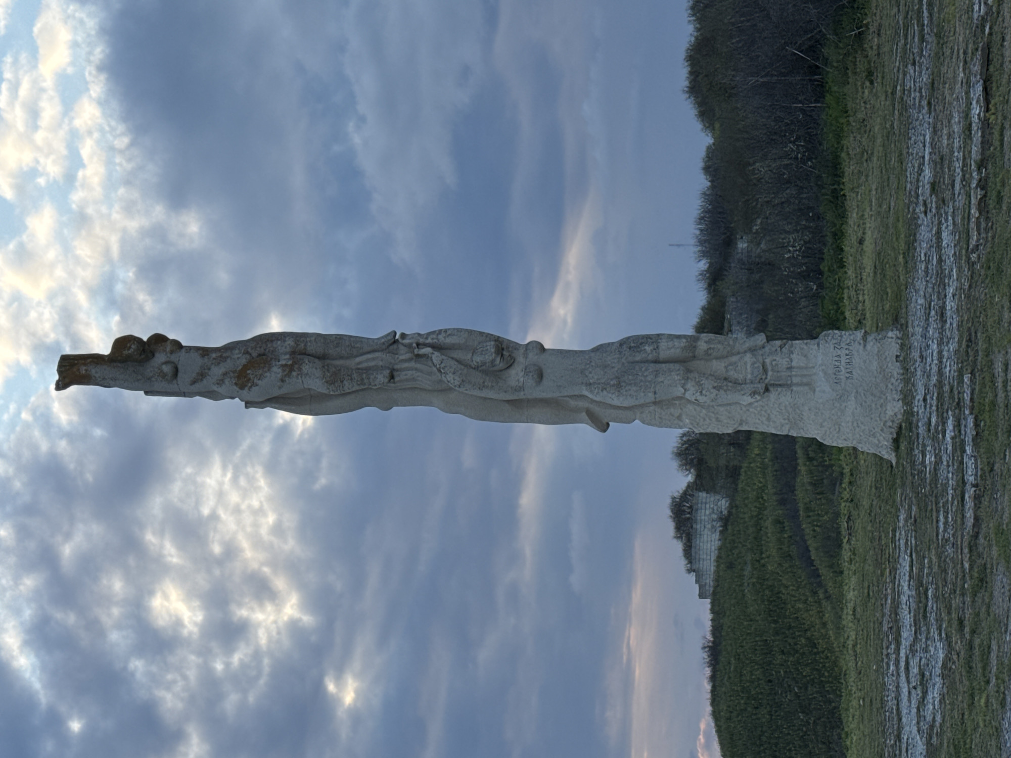 A low-angle, outdoor shot depicts a light-colored, tall, stone monument against a cloudy sky in Balgarevo, Bulgaria. The monument features a series of carved figures and some text in Bulgarian on its base. The vegetation and stone structure in the background suggest the monument is situated in an open field or grassy area near a hill. The monument appears to be a memorial or sculpture of significance, possibly commemorating a historical event or figures associated with the location.