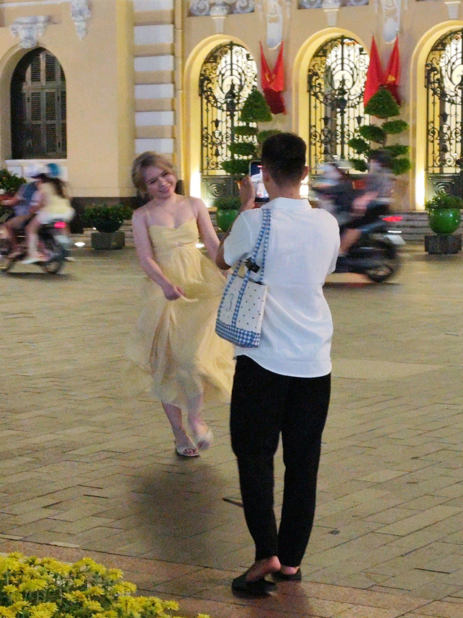 The image depicts a street scene in Ho Chi Minh City, Vietnam, likely in front of a colonial-era building characterized by arched windows and ornate architectural details. In the foreground, a young woman with blonde hair, wearing a flowing yellow dress, is captured mid-twirl, her dress billowing around her. She is smiling and looking towards the camera. Behind her, a person, seen from the back, is holding up a smartphone to take a photograph of the woman. This individual is wearing a white t-shirt and black pants, and carrying a blue and white checkered tote bag over their shoulder. The ground is paved with rectangular stones arranged in a herringbone pattern. In the background, several motorbikes are visible, indicating the common mode of transportation in the city. Red flags and decorative green foliage add to the festive or formal atmosphere. The lighting suggests it is either late afternoon or evening, with artificial lights illuminating the scene. In the bottom left corner, a patch of yellow flowers is visible.