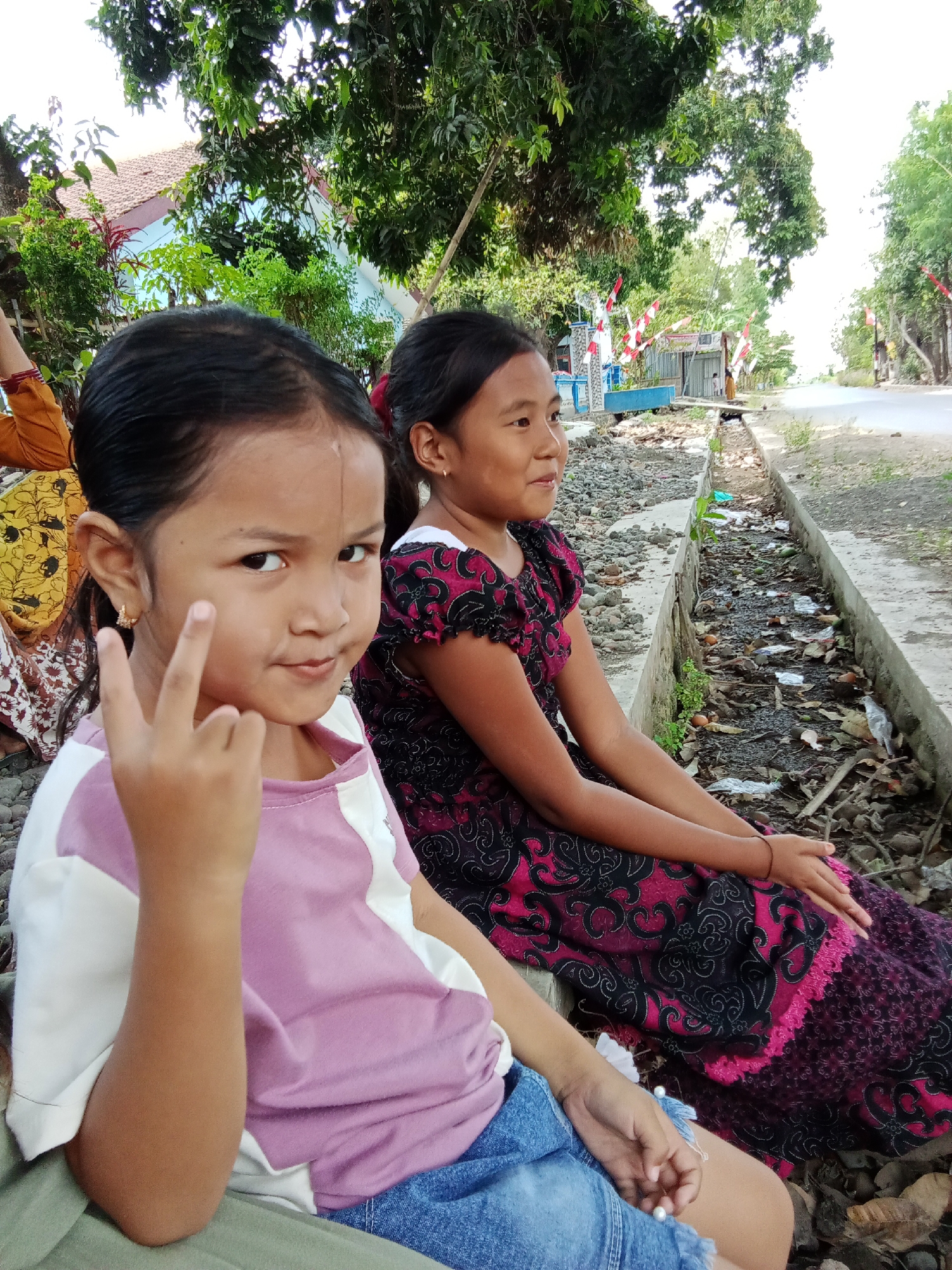 In Trebungan, Indonesia, two young girls are seated outdoors on a concrete structure next to a drainage ditch. The girl in the foreground, facing the camera, is wearing a pink and white t-shirt and blue jeans. She is making a peace sign with her right hand. The girl behind her is wearing a dark dress with a pink floral pattern and is looking to her right. The drainage ditch is filled with debris, including leaves, plastic, and stones. In the background, trees line a road, and there are houses and small structures visible. Red and white flags are strung along the roadside.