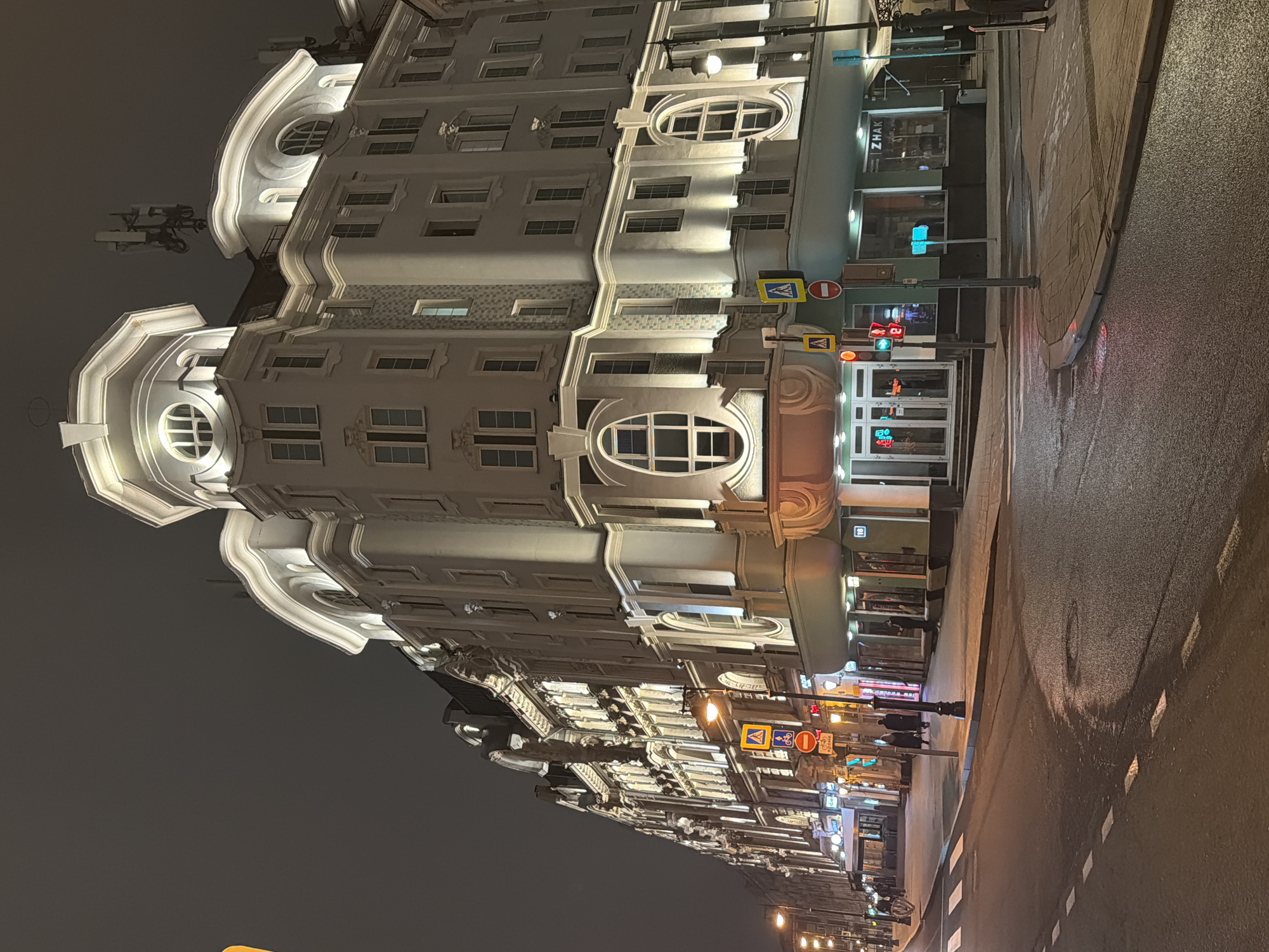 The image depicts a street scene in Moscow, Russia, at night. The focus is a multi-story building with ornate, light-colored architecture. The building's façade includes numerous windows, decorative moldings, and illuminated oval features. Streetlights and signage are visible along the street. Traffic lights and pedestrian crossing signs are present. The street surface appears wet. The lighting suggests an urban environment at dusk or nighttime. There are a few pedestrians in the distance, but no visible human interactions.