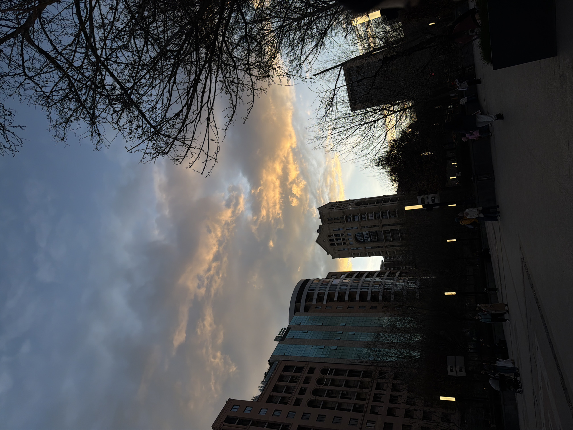 The image shows a street in Yerevan, Armenia, at dusk. Tall, modern buildings line one side of the street, partially obscured by bare trees. The sky is filled with clouds, some tinged with orange from the setting sun. People walk along the sidewalk. Streetlights illuminate the scene.