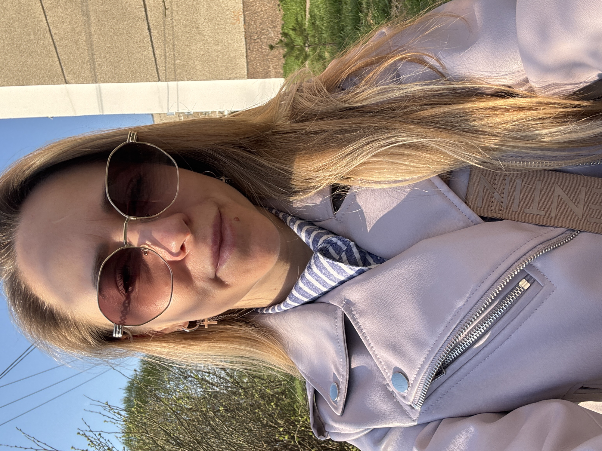 A selfie of a woman wearing sunglasses and a light purple leather jacket over a blue and white striped shirt. She is wearing a necklace with a cross pendant. The background shows a building and green trees, suggesting an outdoor location. The image could be taken in an urban area, possibly the city of Chaikovsky, Russia, but the specific location is not identifiable within the provided image.