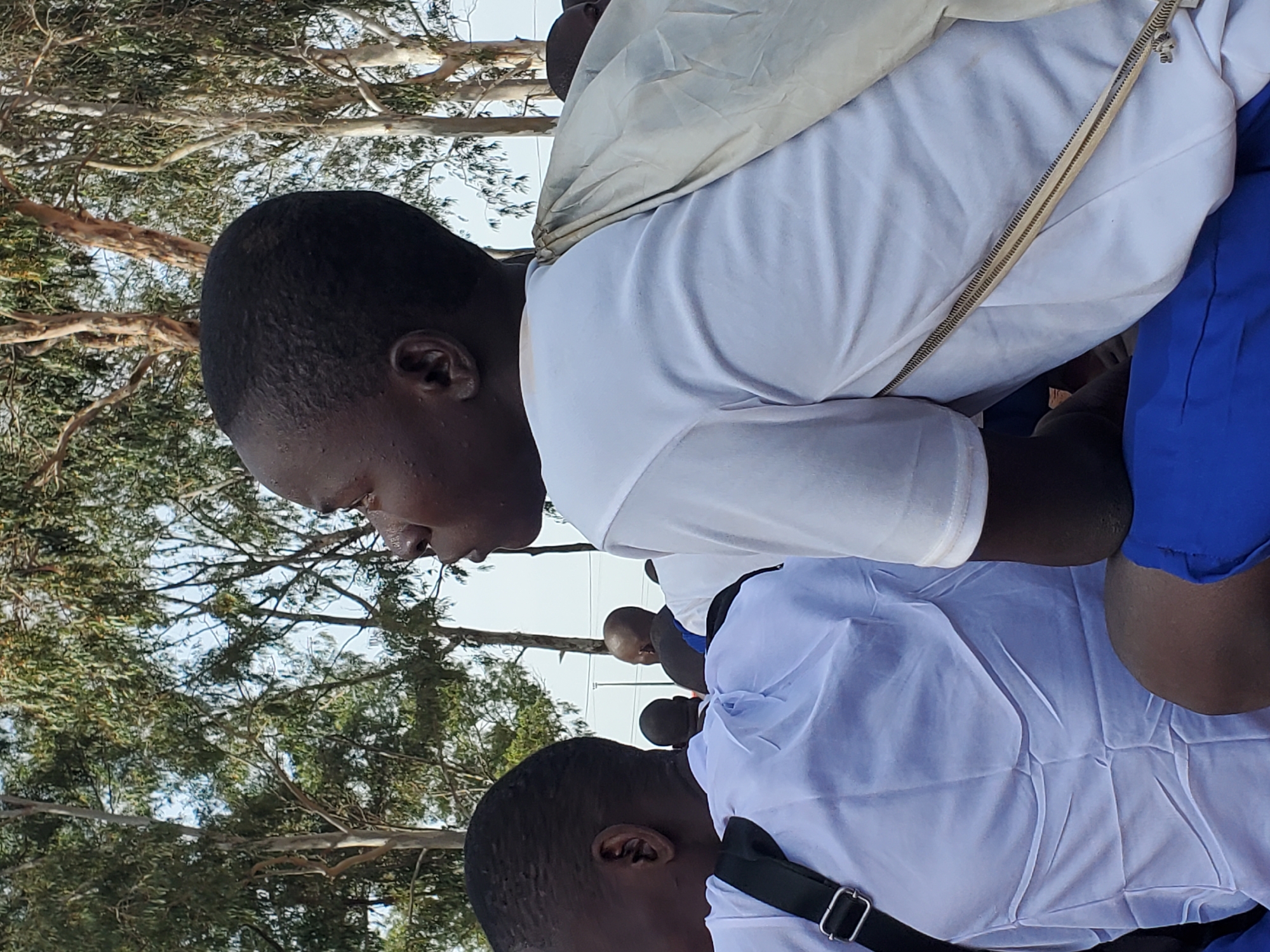 The image shows a medium shot of a group of young people, likely students, from the chest up, seated outdoors. The main subject, a young person with dark skin and short hair, is positioned in the foreground, facing to the right. They are wearing a white t-shirt with blue shorts. A light-colored cloth is draped over their left shoulder, fastened with a zipper-like detail. Behind them, other individuals are also visible, mostly wearing white tops. The background consists of tall trees with sparse foliage, suggesting a rural or semi-rural environment, possibly within Barakin Rafin Gora, Nigeria. The lighting indicates it is daytime, with the sun casting soft shadows. The overall atmosphere appears calm and focused, as if the subjects are observing something or waiting for an event.