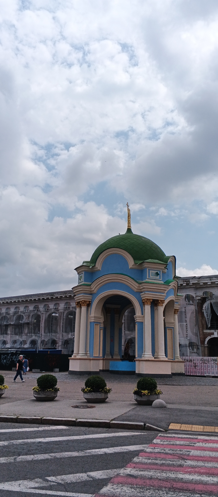 The image captures a street scene in Kyiv, Ukraine, featuring a small, ornate blue and white pavilion with a green dome topped by a golden cross. The pavilion is situated in the foreground, with arched openings revealing a seated figure inside. To the left of the pavilion, a building is partially visible, draped in scaffolding and sheeting with text printed on it, suggesting ongoing construction or renovation. In front of the pavilion, a pedestrian crossing with white and pink stripes is visible. Several round planters with green bushes line the pavement. Two individuals are seen walking on the pavement to the left of the pavilion. The sky above is filled with scattered clouds.