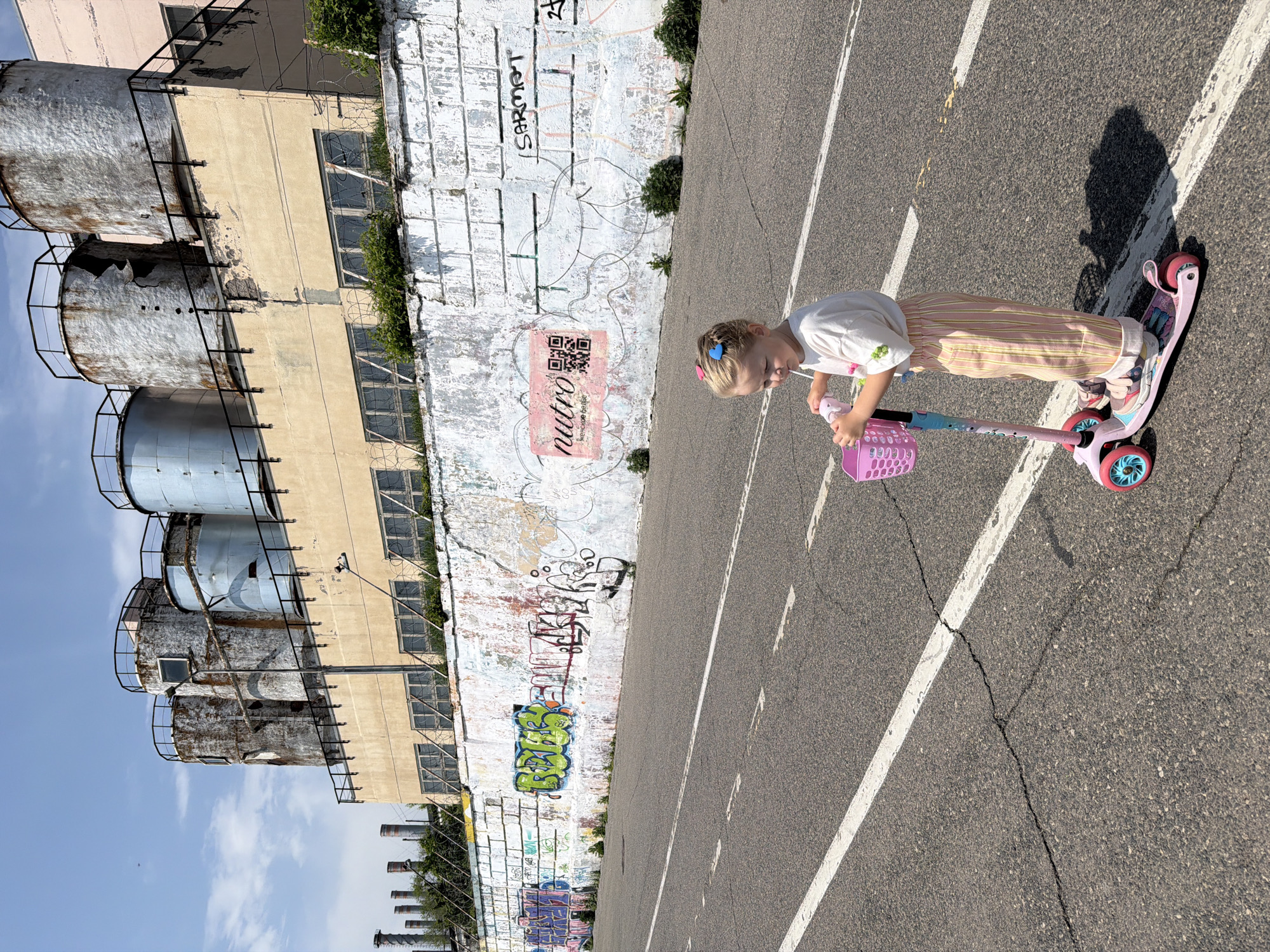 A young girl rides a pink scooter on an asphalt surface marked with white lines. She wears a white shirt and striped pants, and has a pink basket attached to the scooter's handlebars. Behind her is a graffiti-covered wall next to an industrial building. The image is likely taken in Saratov, Russia, based on wall markings visible.