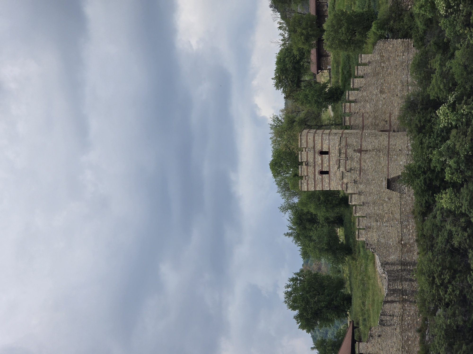The image shows a stone fortress with a tower, possibly part of a castle, with a cloudy sky in the background. The fortress is made of stone blocks, with a tower featuring narrow openings. It is partially surrounded by trees and green vegetation. The fortress appears old and weathered. The photo suggests a historical site in a natural setting.