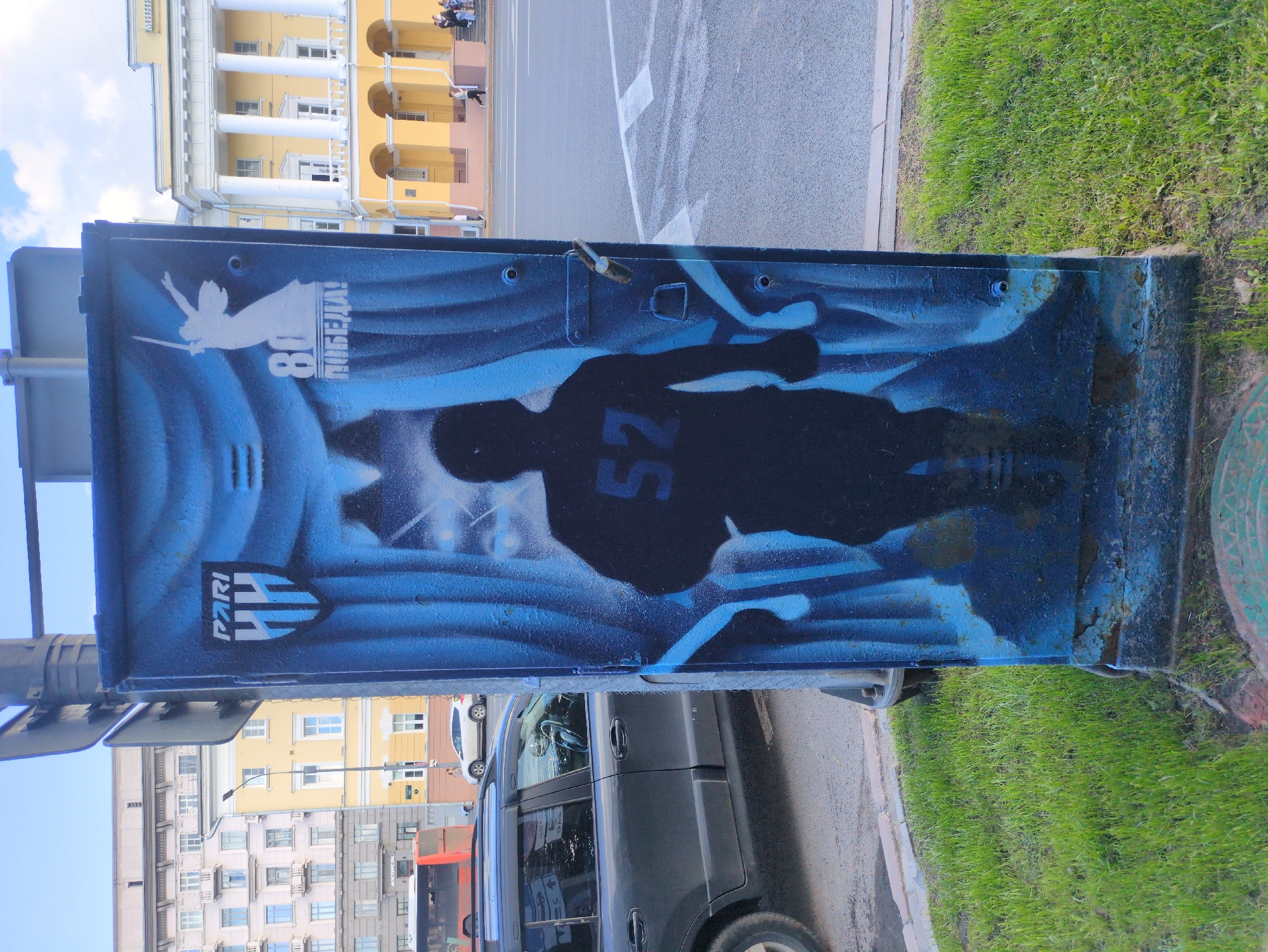 This media file features a close-up of a graffiti-covered metal structure, likely a utility box or a portion of a street fixture. The graffiti is primarily in shades of blue and black, depicting a silhouette of a person in athletic gear, with the number "52" prominently displayed on their chest. To the left of the silhouette, there's a white outline of a winged figure holding a sword, and the number "80" next to the Russian word "ПОБЕДА!" (Pobeda!, meaning "Victory!"). Below the silhouette, a shield-like logo with the word "PARI" and what appears to be stylized letters "HH" is visible.
In the background, a yellow building with classical architectural elements, including columns, is seen. Further back, there are other buildings of varying colors and architectural styles, suggesting an urban environment. A street with white markings and a sidewalk bordering a patch of green grass are also present. A dark-colored van and a red bus are partially visible in the lower portion of the image, indicating ongoing traffic. The sky is a bright blue, suggesting it's daytime with clear weather. The overall scene is static, focusing on the graffiti art and its urban backdrop in Nizhny Novgorod, Russia.