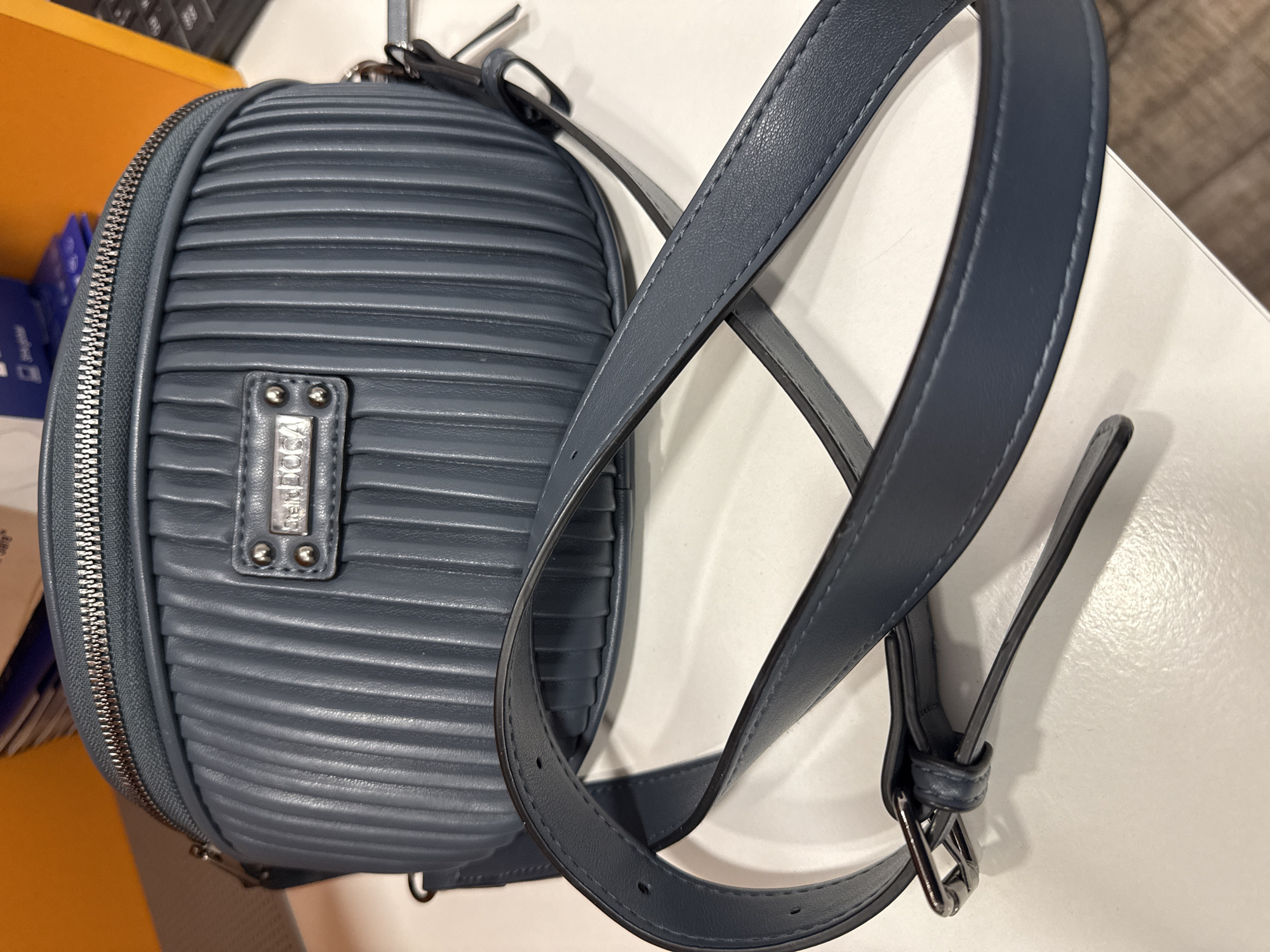 A blue leather backpack is on a white surface. The backpack has a zipper closure and a metal nameplate. The shoulder straps are blue leather with adjustable buckles. The background is out of focus and shows a partial view of a keyboard and yellow object.