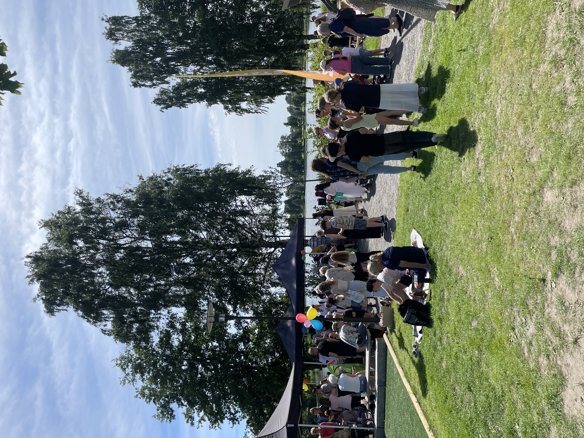 The image depicts a sunny day event taking place outdoors in Karlstad, Sweden. A crowd of people are gathered, suggesting a festival, market, or social event. Tents are set up, likely for vendors or shade, and a yellow banner is visible in the background. Balloons, including red, blue, and yellow, add a festive touch. People are dressed in casual summer attire. The setting appears to be a park or recreational area with lush green grass and trees. The sky is partly cloudy, indicating fair weather, and the shadows suggest it is midday. The overall mood seems relaxed and social. There is no visible text in the image.