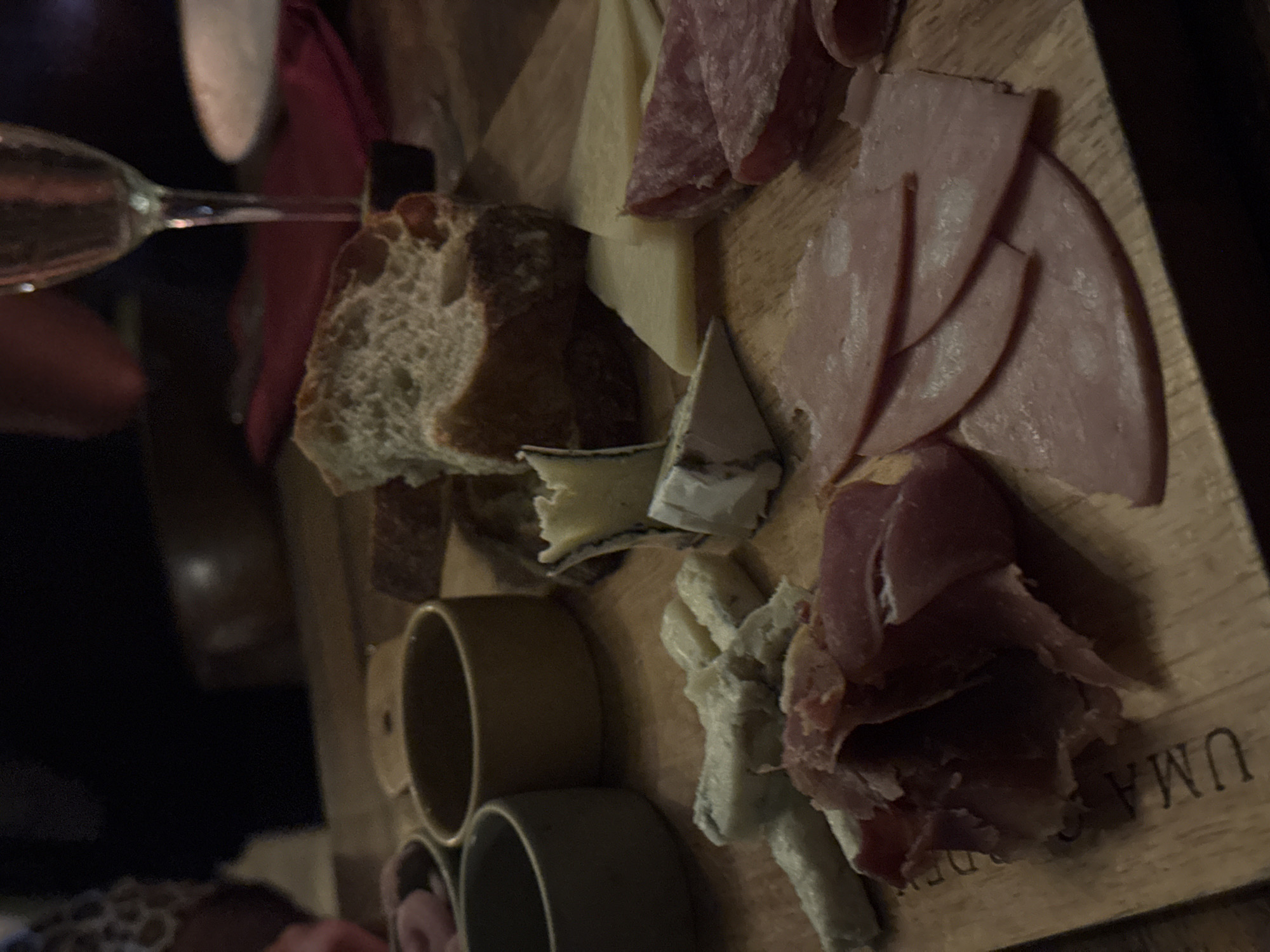 This is a close-up, medium shot of a charcuterie board on a wooden surface, likely in North Kuta, Indonesia. The board is laden with various cured meats and cheeses, arranged artfully. To the left, two small, dark-colored bowls are visible, one containing pinkish items, possibly olives or cherry tomatoes. In the center, a rustic loaf of bread is partially visible, with its crusty exterior and airy interior exposed. Several types of cheese are present, including a crumbly blue cheese, a wedge of brie or camembert with a rind, and slices of hard yellow cheese. Slices of red salami and pink ham are fanned out across the board. In the background, a glass of white wine and a red napkin are slightly out of focus. The lighting suggests an indoor setting, likely a restaurant or bar, contributing to a cozy, intimate ambiance. The overall scene depicts a culinary offering, emphasizing the textures and colors of the food.