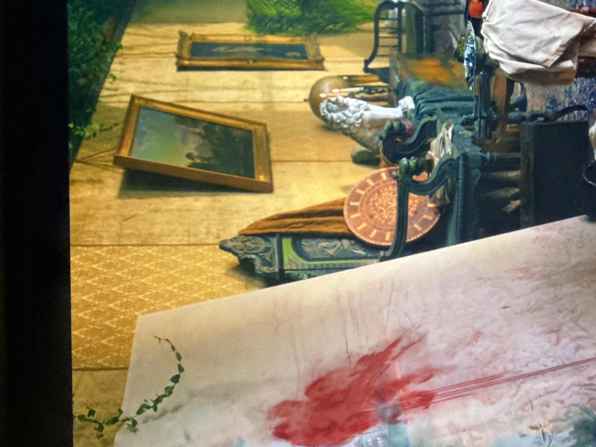 The image shows a room interior in Puebla, Mexico. On a patterned tan surface, there are two framed landscape paintings. A white bust and a decorative plate rest near a dark green ornate object. A painter's canvas, marked with a red smear, takes up the foreground. A tendril of greenery grows from the edge of the canvas. An elaborate dark machine sits to the right.