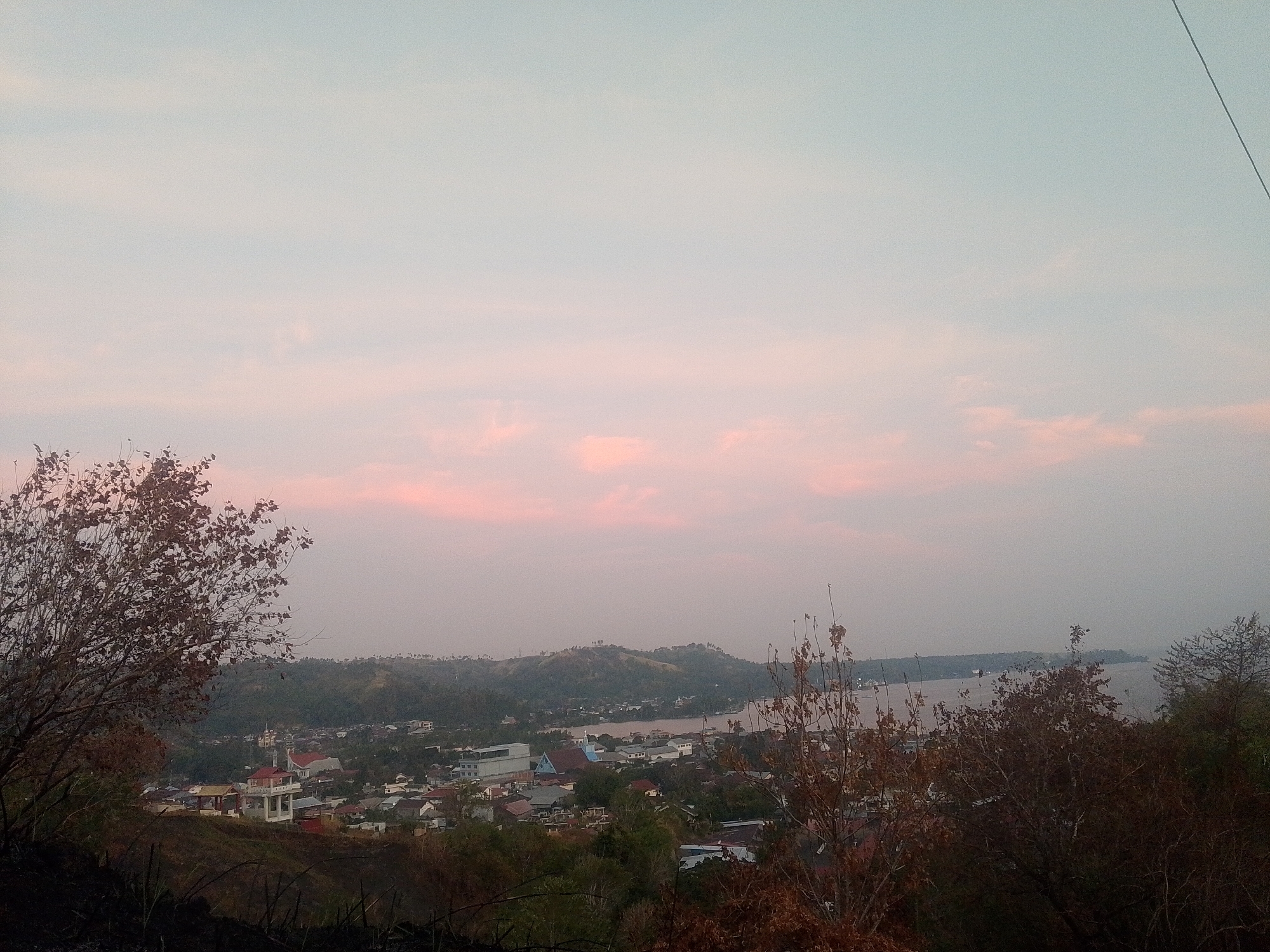 The image presents a wide, elevated view of a coastal town at dusk. The sky is filled with soft, pastel colors of pink and light blue, indicating either sunrise or sunset, with scattered wispy clouds. In the foreground, the silhouettes of dry, leafless trees and burnt vegetation are visible on a dark, sloping hillside.
Further down the slope, a town with red-tiled roofs and white buildings spreads out. Several structures are clearly distinguishable, including a prominent building with a red roof and white supporting pillars, and a church with a white steeple and a red roof. The town is nestled amongst rolling green hills.
Beyond the town, a body of water, likely a bay or strait, stretches to the horizon. A thin strip of land or a distant island with trees can be seen across the water. The overall atmosphere is serene and tranquil, with a sense of quiet settlement overlooking the water.
The location is identified as Buyungon, Indonesia.
