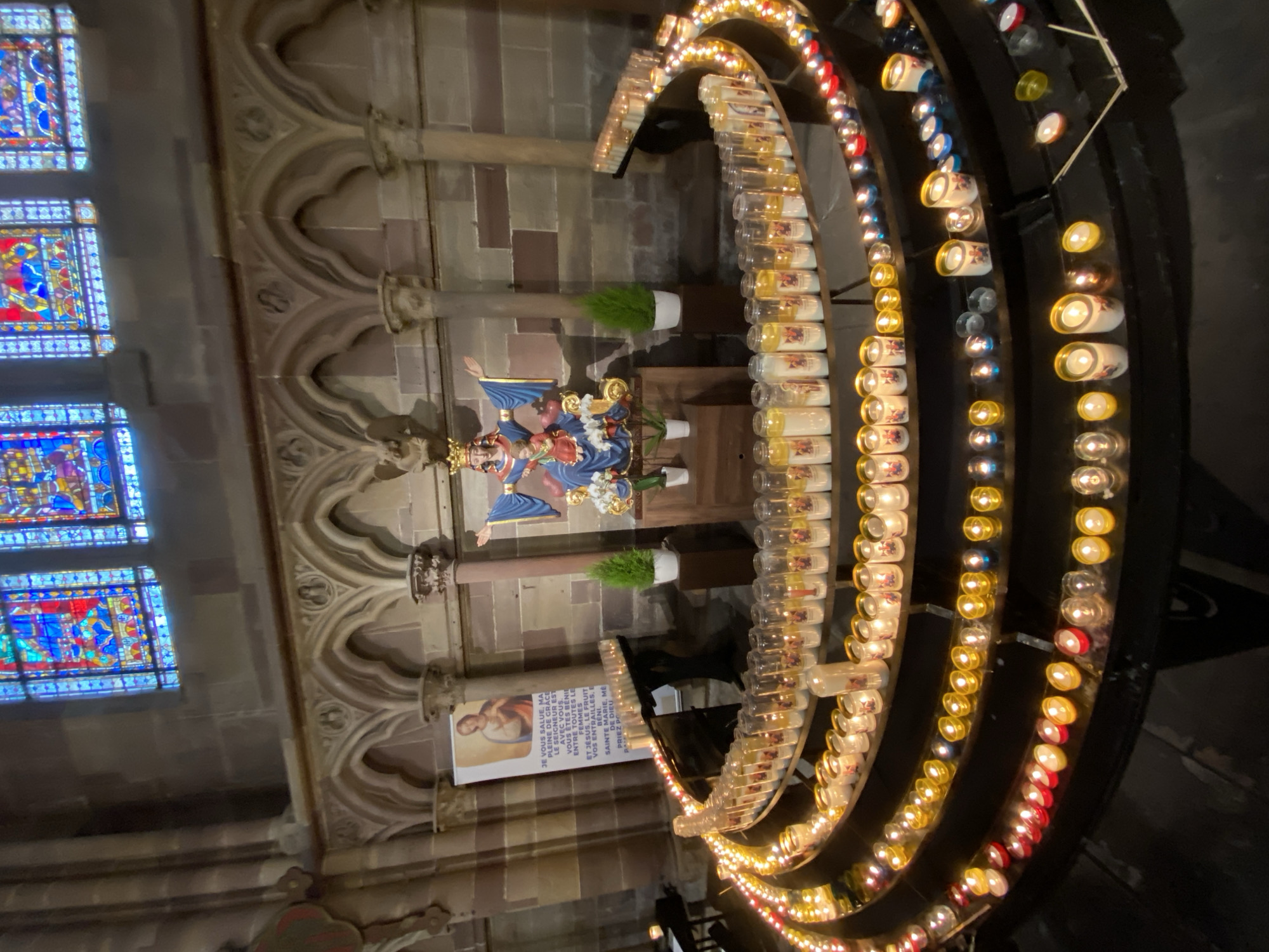 The image, oriented vertically, displays the interior of a church in Strasbourg, France. A central religious statue depicts the Madonna and Child, adorned in blue, gold, and red garments, with the Virgin Mary wearing a crown. The statue rests on a pedestal against a textured stone wall featuring elaborate arched details and columns.
To the right of the statue, a large, multi-tiered candelabra or votive candle holder extends in a graceful curve, illuminating the scene with numerous lit candles. Many candles are enclosed in clear or decorated glass jars, while others burn openly, displaying a spectrum of colors including yellow, red, and white. On the left, three vibrant stained glass windows with intricate blue, red, and yellow patterns provide additional illumination and rich color.
The setting reveals the grand architecture of a cathedral, characterized by detailed stone carvings and robust structural elements. Two small potted green plants flank the statue's base. A sign or poster is visible to the left of the candelabra, displaying text in French, which reads in part: "JE VOUS SALUE, MA PLEINE DE GRACE LE SEIGNEUR EST AVEC VOUS. VOUS ÊTES BÉNIE ENTRE TOUTES LE FEMMES ET JÉSUS, LE FRUIT VOS ENTRAILLES, E BENI DE DIEU SAINTE MARIE, ME PRIEZ PO," a section of the "Hail Mary" prayer. The atmosphere is serene and devotional, characteristic of a sacred space.