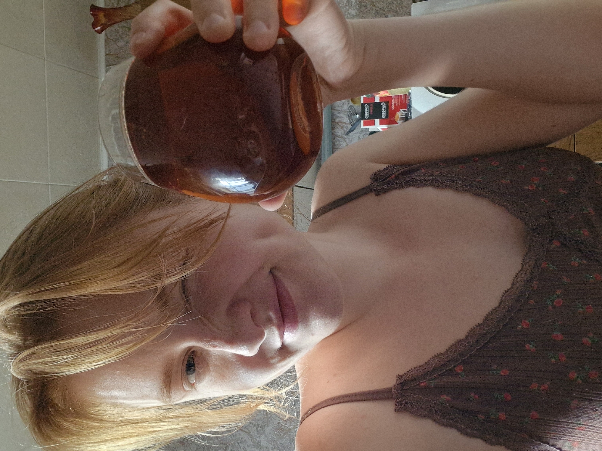 A woman is holding a glass jar filled with a dark brown liquid, likely tea. She's in an indoor setting, possibly a kitchen, with tiled walls and a counter visible in the background. Boxes of Cheez-It crackers are visible on the counter. The woman has light skin, reddish-blonde hair, and is wearing a brown floral top. The image has been rotated. There is no clear indication of a location in the city of Yekaterinburg, Russia.
