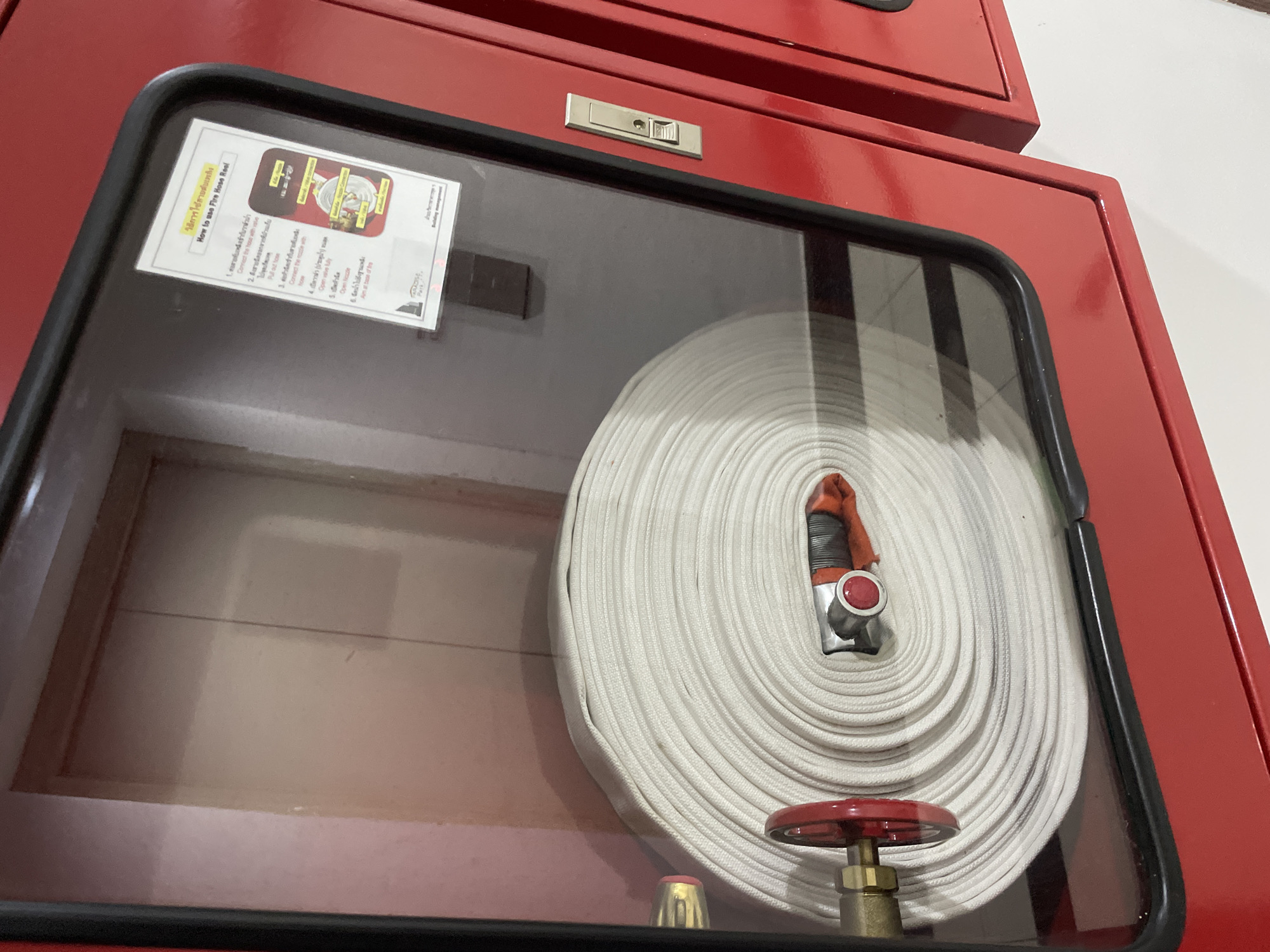 The image shows a red fire hose cabinet with a glass door, revealing a tightly coiled white fire hose inside. A red valve wheel is visible on the left side of the hose reel. On the inside of the glass door, a notice with instructions on "How to use Fire Hose Reel" is affixed. The notice has text in both English and Thai, with illustrations depicting the steps. In the background, through the glass, a door with a light pinkish hue can be seen. A silver latch is present on the right side of the cabinet door. The setting appears to be an indoor environment, possibly a building common area or hallway. The style of the cabinet and the fire safety equipment suggest it is a public or commercial building.