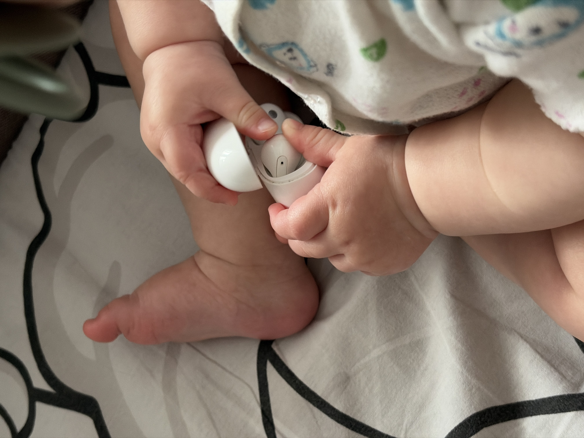 A baby is seated on a black-and-white patterned sheet, likely in a crib or bassinet. The baby is wearing a white onesie with light-colored patterns. The baby is holding a white, open case of wireless earbuds in their hands, and at least one earbud is visible inside the case. The baby's foot is visible on the left side of the image. The location and the time of day can not be determined with this image.