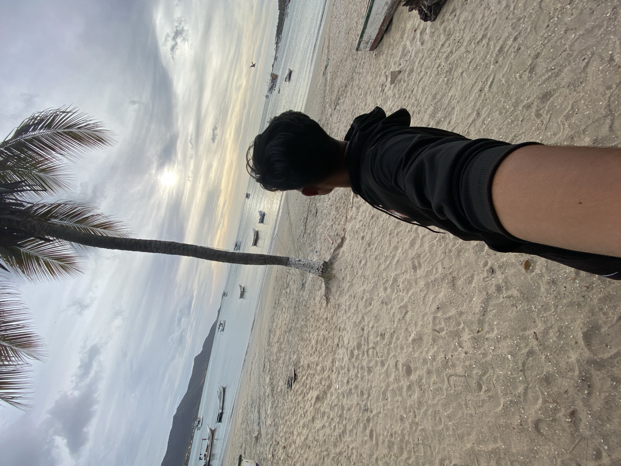 The image presents a coastal scene in Juan Griego, Venezuela, observed from a first-person perspective on a sandy beach. A single individual, with dark hair and wearing a black hooded sweatshirt, is positioned in the lower right and center, viewed from behind. The individual's right arm is extended forward, holding what is presumed to be the camera, and they are facing the bay.
The foreground and midground consist of light-colored sand with visible ripples and scattered small shell fragments. A tall palm tree, its base partially painted white, stands on the beach slightly to the right of the frame's center, extending upwards with its fronds visible against the sky. To the far right, a section of a white and green wooden boat, possibly beached or derelict, is visible on the sand, adjacent to dark, weathered wooden debris.
The calm waters of the bay occupy the midground, reflecting the overcast sky. Approximately a dozen small fishing boats, mostly white or light-colored, are anchored at various distances across the bay. In the distance, a range of dark, undulating mountains forms the horizon line. The sky is predominantly overcast with layers of grey clouds, though a bright, diffused sun is visible through a break in the clouds in the upper right quadrant. A dark bird is in flight against the sky in the upper mid-right.