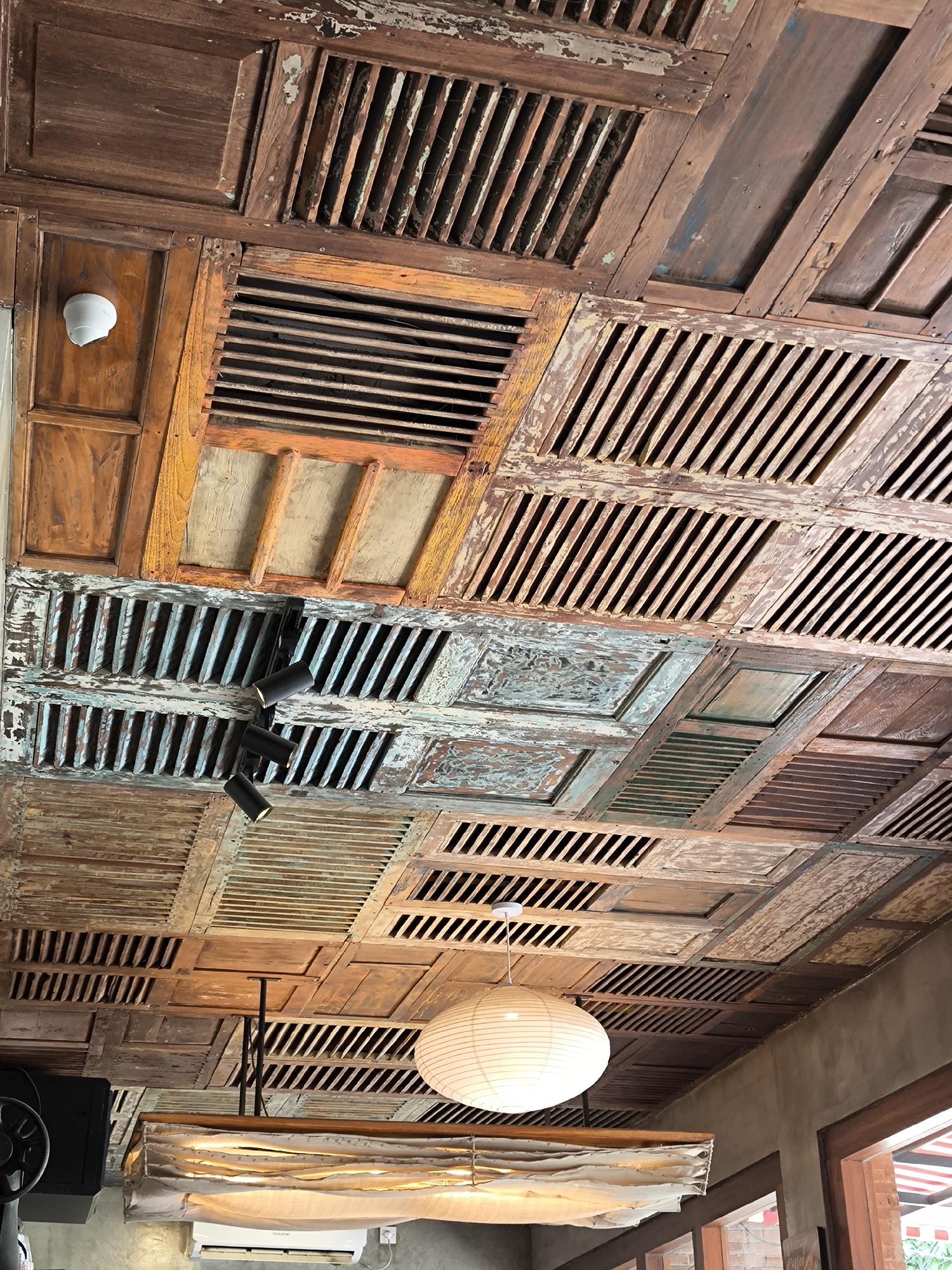 The image shows a ceiling constructed from salvaged or repurposed wooden window shutters in a space, likely a restaurant or cafe, in Unknown, Indonesia. The shutters are arranged in a grid pattern, with varying colors and states of weathering. Some are stained or painted in shades of brown, orange, and blue, while others show signs of paint peeling or general wear. The shutters are mostly in a louvered configuration, but some are solid panels.
Several light fixtures are suspended from the ceiling. One is a modern, rectangular fixture covered with a fabric diffuser, and another is a spherical paper lantern. Track lighting fixtures are also visible, mounted directly onto the shutters. The overall aesthetic is rustic and eclectic.